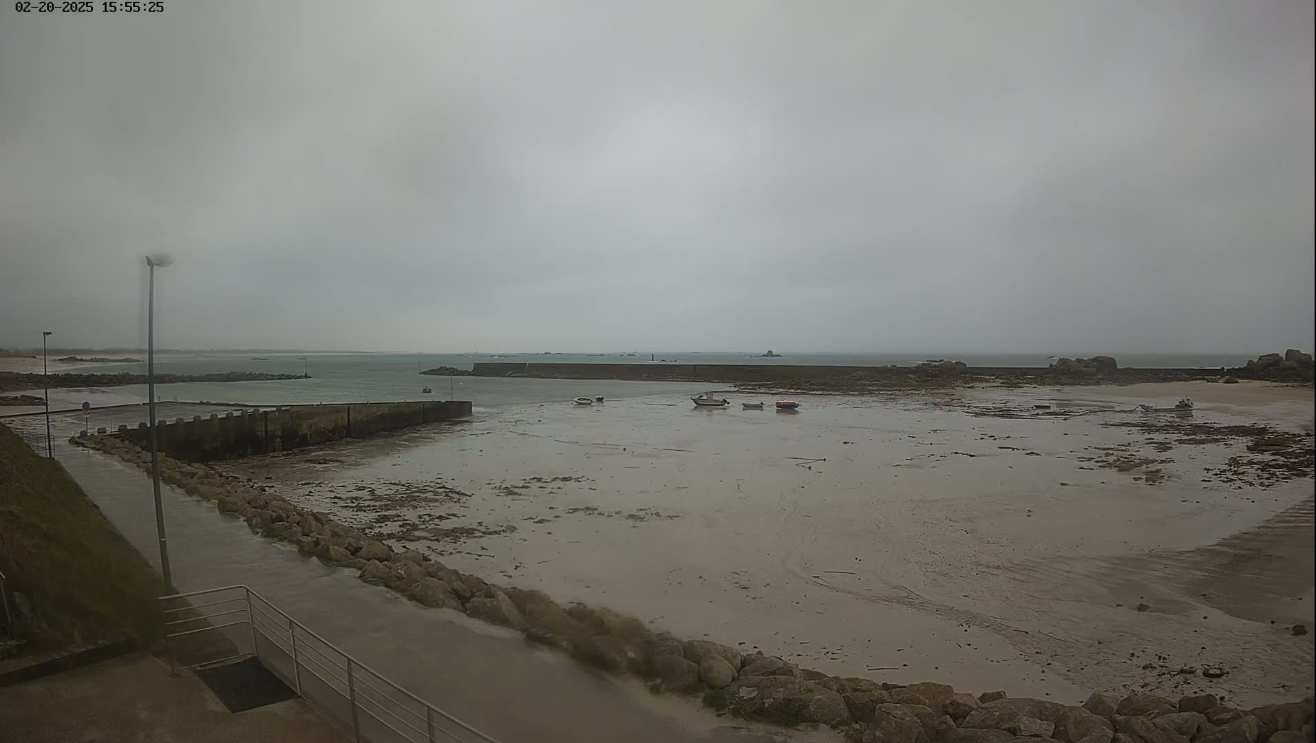 A mostly cloudy day shows a rocky, low-tide beach with several small boats near a harbor wall and calm water extending to a distant shoreline.