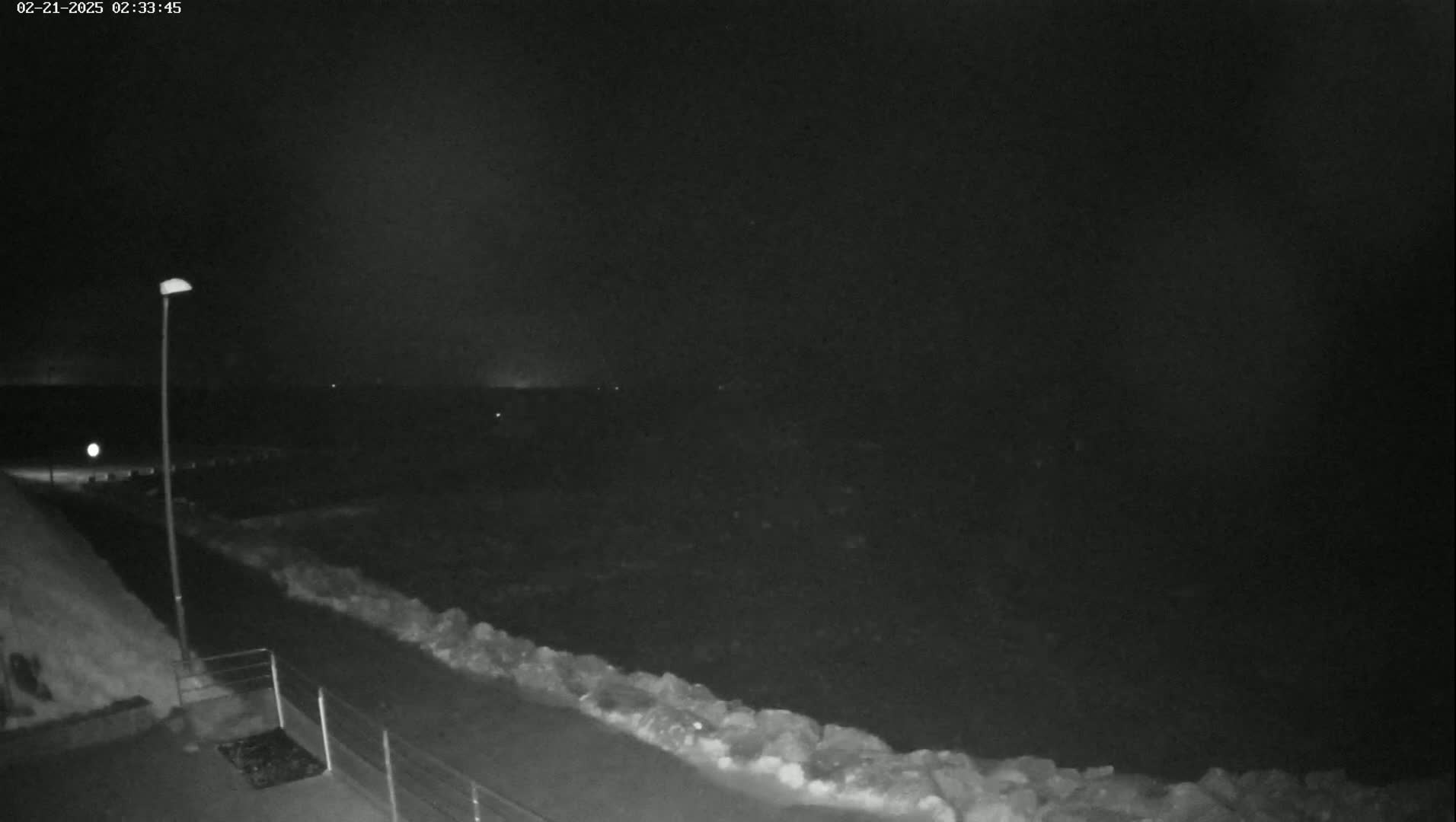 A nighttime view of a paved walkway next to a rocky shoreline, under a dark, possibly overcast sky.
