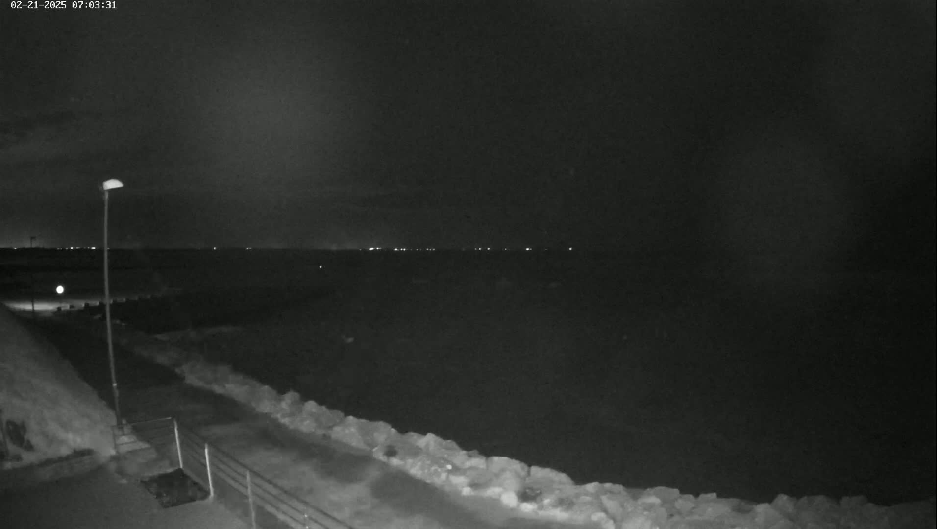 A nighttime view shows a dark body of water with distant lights on the horizon, a paved path next to a low stone wall, and a single streetlight.