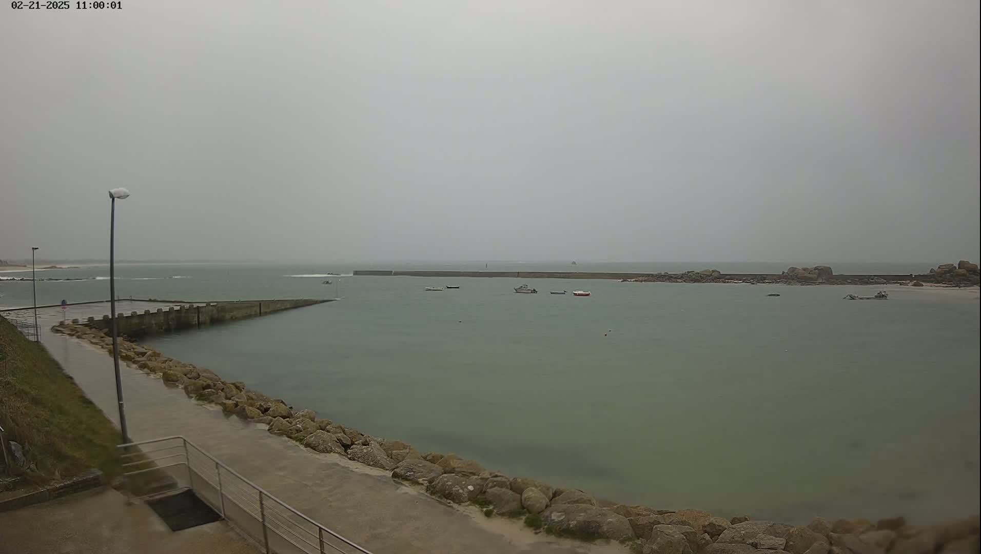 A calm, overcast day shows a harbor with several small boats, a long seawall, and a rocky shoreline.