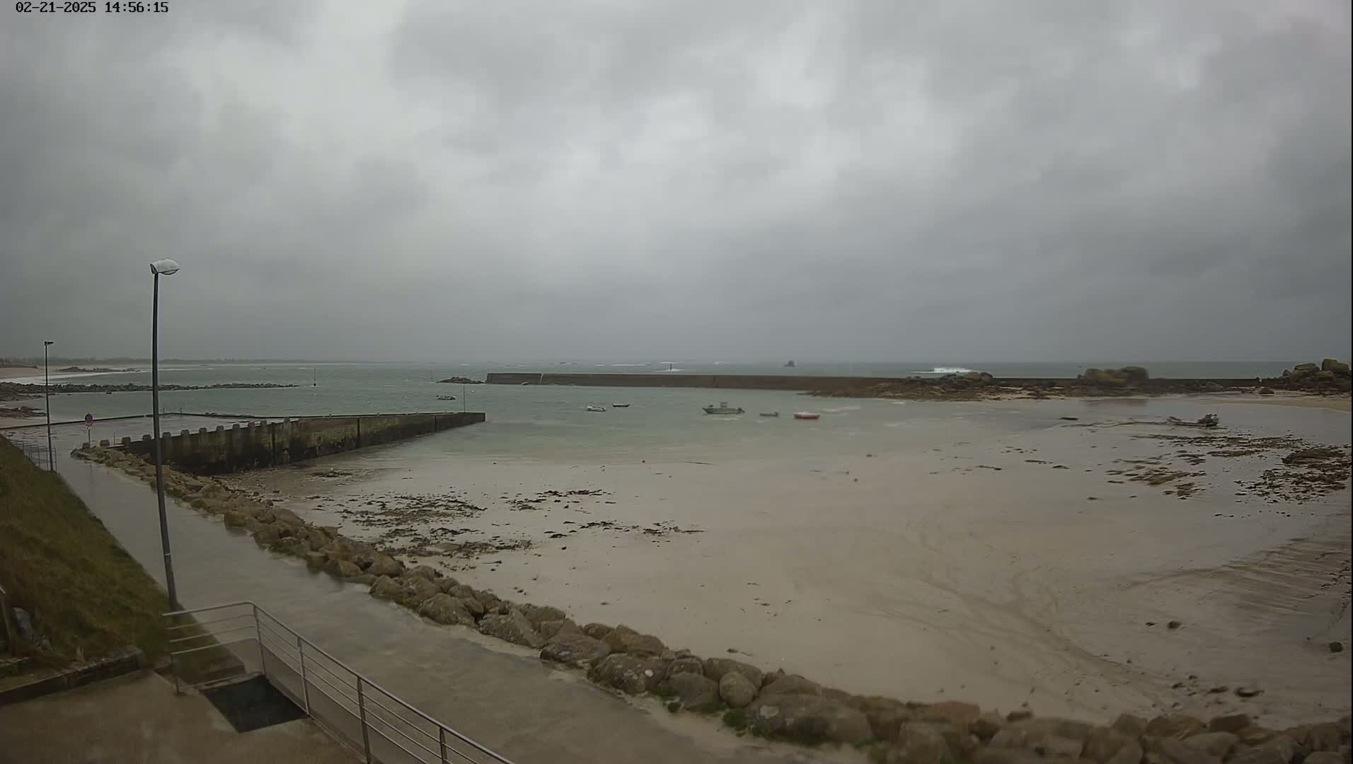 A mostly cloudy day shows a small harbor with several small boats at low tide, a wet walkway leading to it, and a calm ocean in the background.
