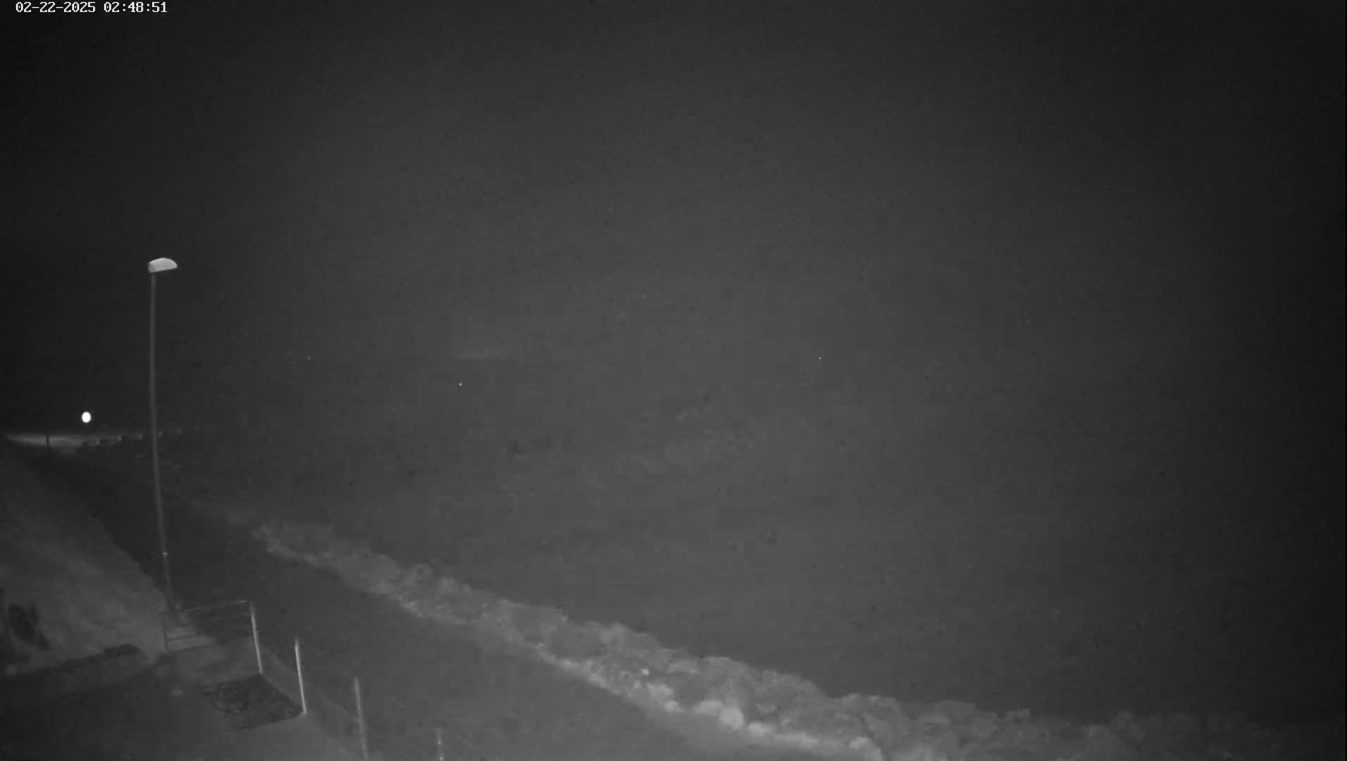 A nighttime, low-light view shows a dark road curving to the right, bordered by a low wall or embankment, with a single lamppost visible to the left.