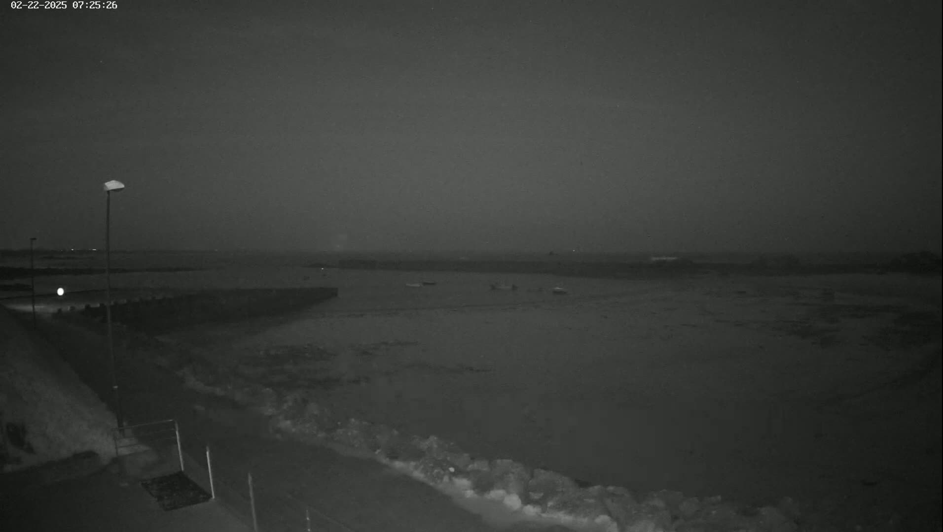 A nighttime, low-light view of a calm body of water, possibly a bay or harbor, with several small boats visible in the distance under a dark, overcast sky.