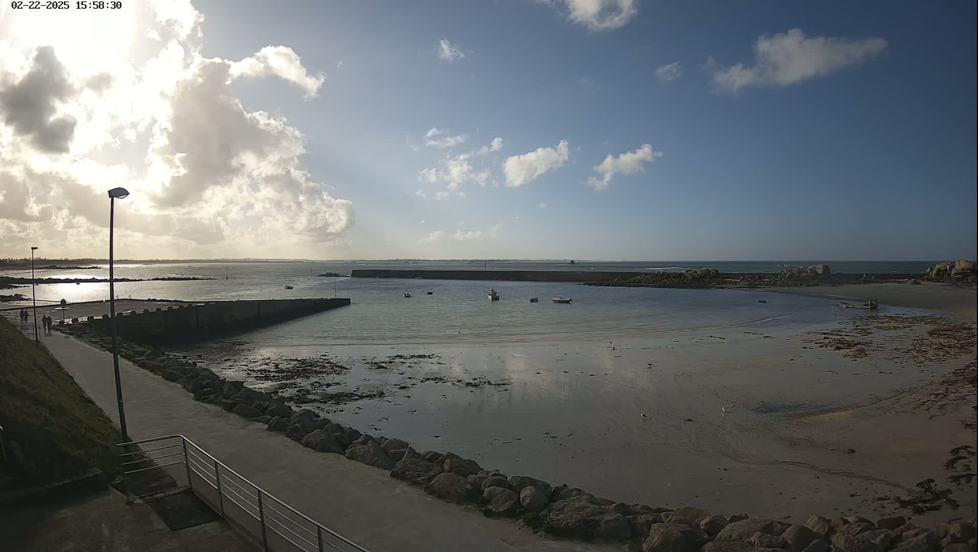 A mostly sunny day shows a harbor with several small boats, a calm sea revealing exposed sand and rocks at low tide, and a paved walkway leading to the water's edge.