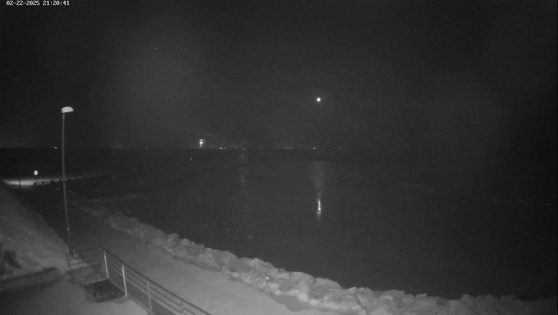 A nighttime, snow-covered waterfront scene shows a body of dark water under a dark sky with a few faint lights in the distance.