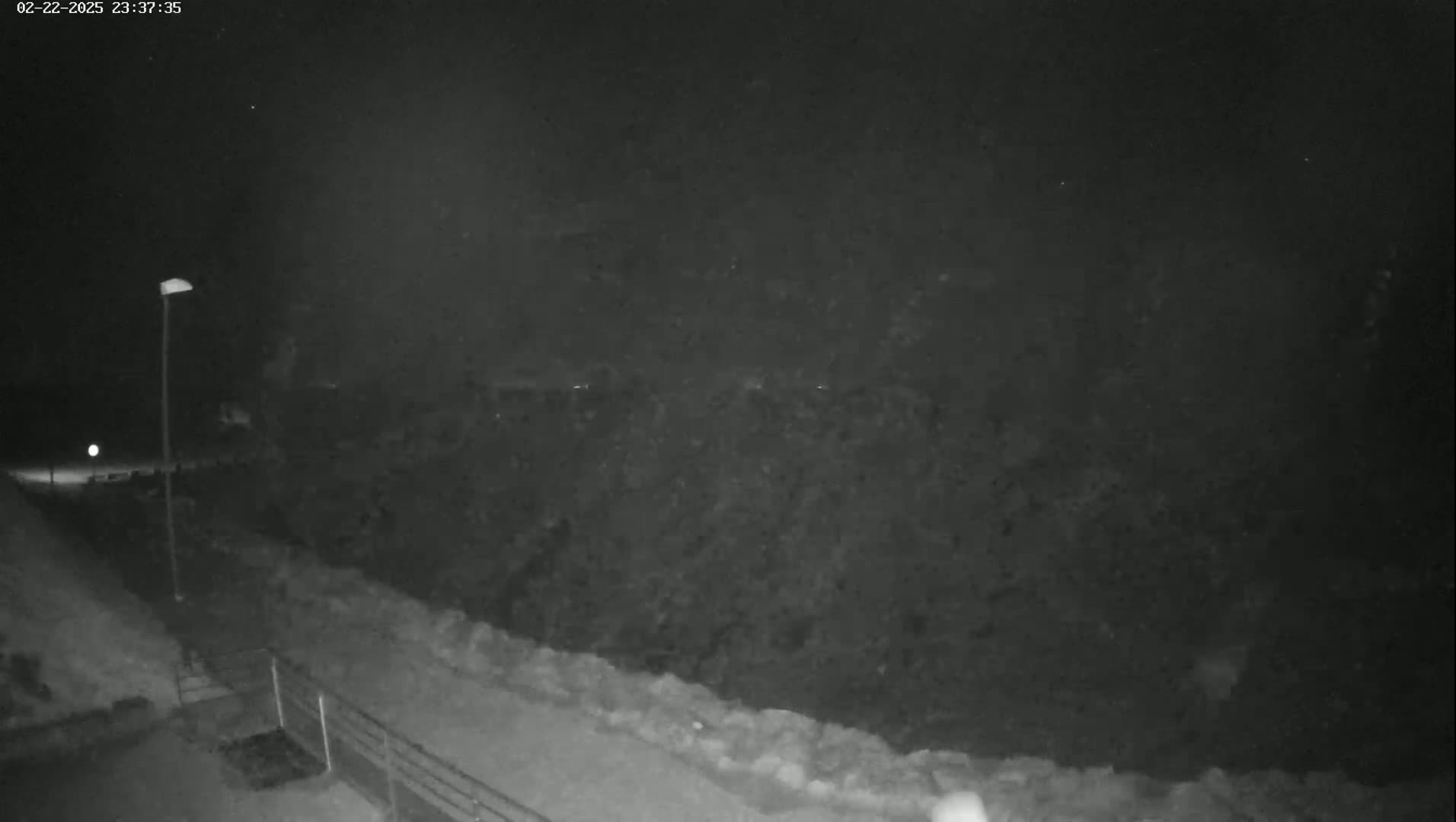 A nighttime, low-light view shows a dark hillside sloping down to a snow-covered path illuminated by a single lamppost.