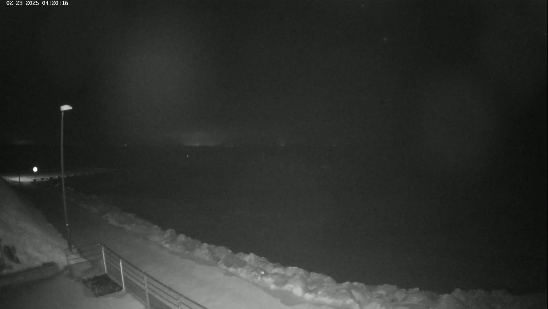 A nighttime, snowy scene shows a snow-covered walkway next to a body of water under a dark sky.