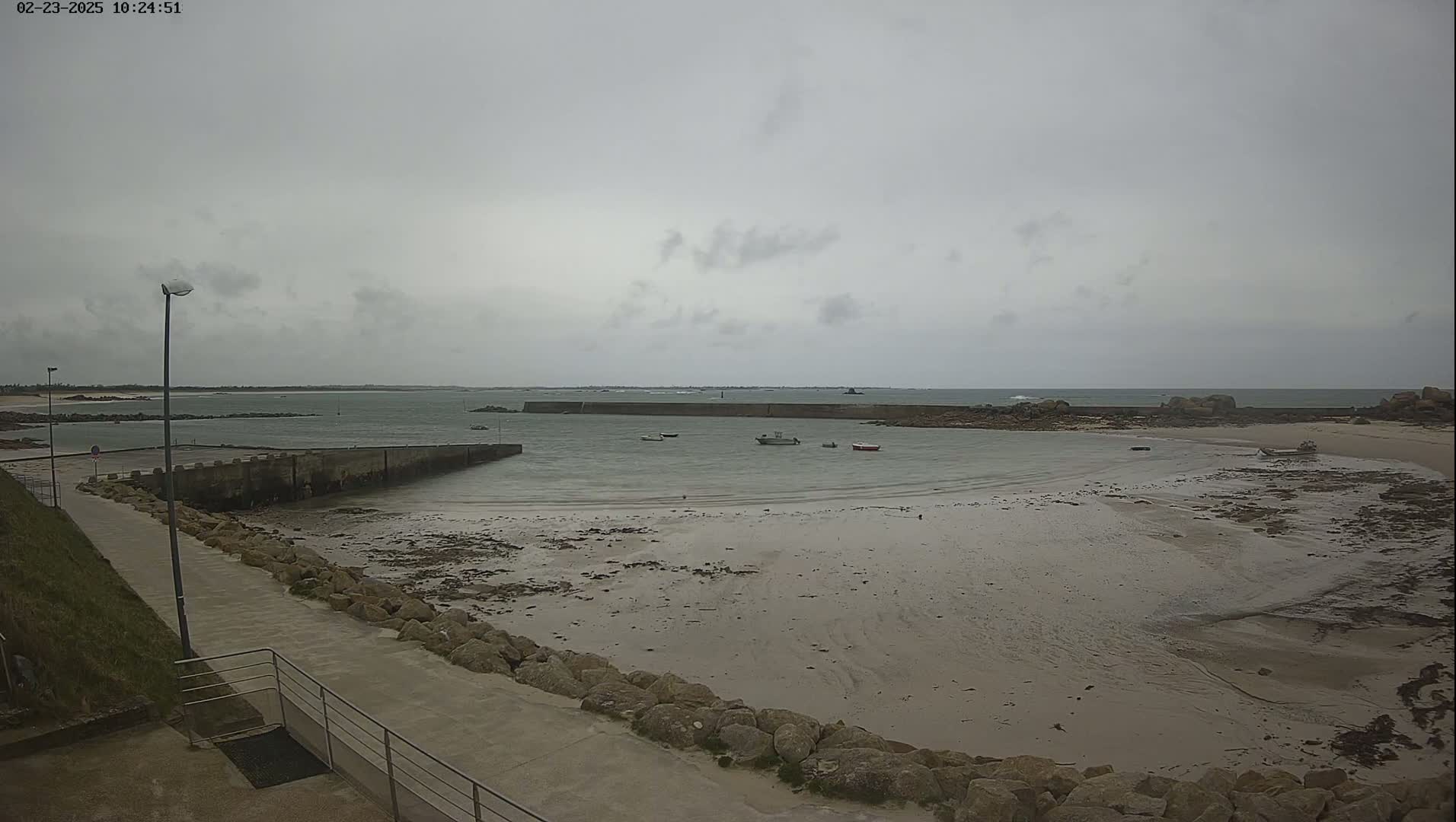 A mostly overcast sky hangs over a calm bay with several small boats moored near a stone jetty and a sandy beach at low tide.