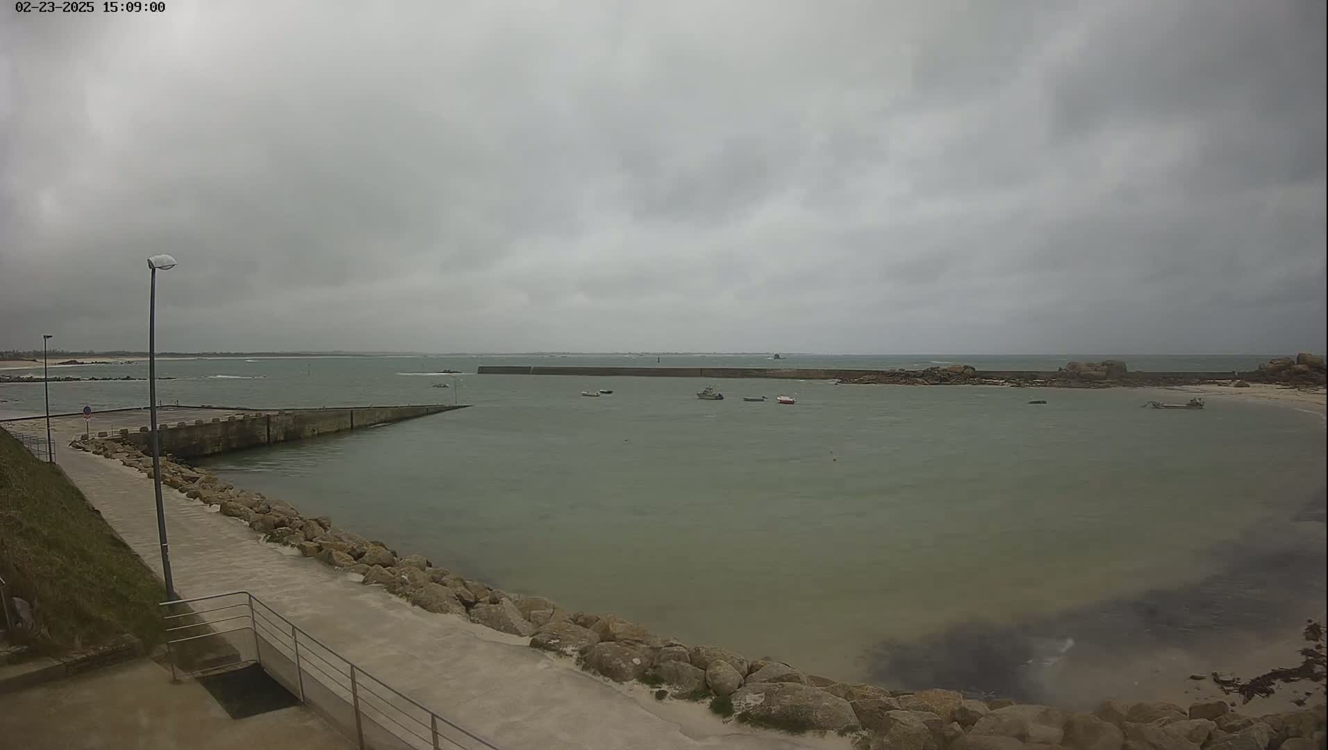 A calm, overcast day reveals a small harbor with a few boats, a stone seawall, and a paved walkway leading to the water.