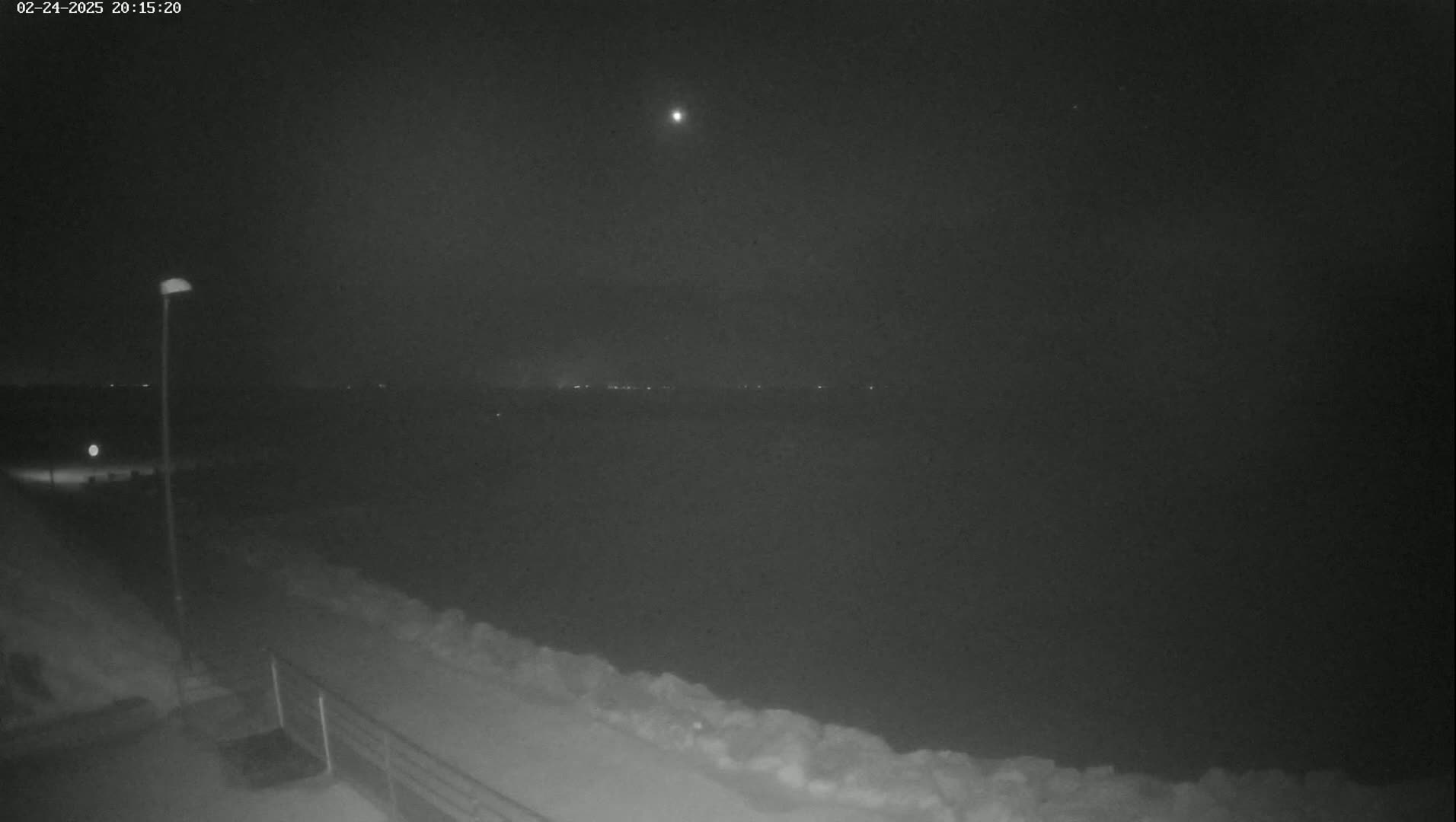 A nighttime view shows a snow-covered landscape with a distant, faintly lit horizon under a dark, mostly clear sky with a bright light, possibly the moon, visible.