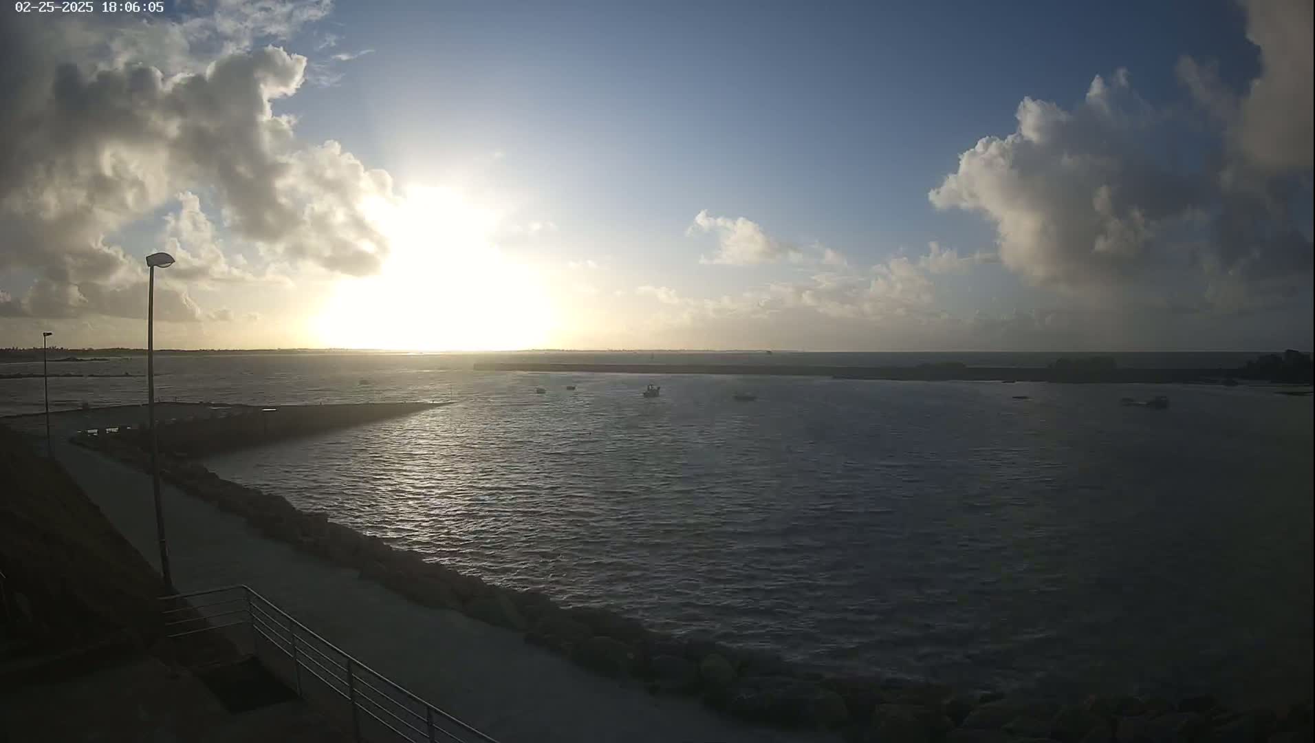A partly cloudy sunset over a calm body of water with a few small boats and a paved walkway beside a rocky shoreline.