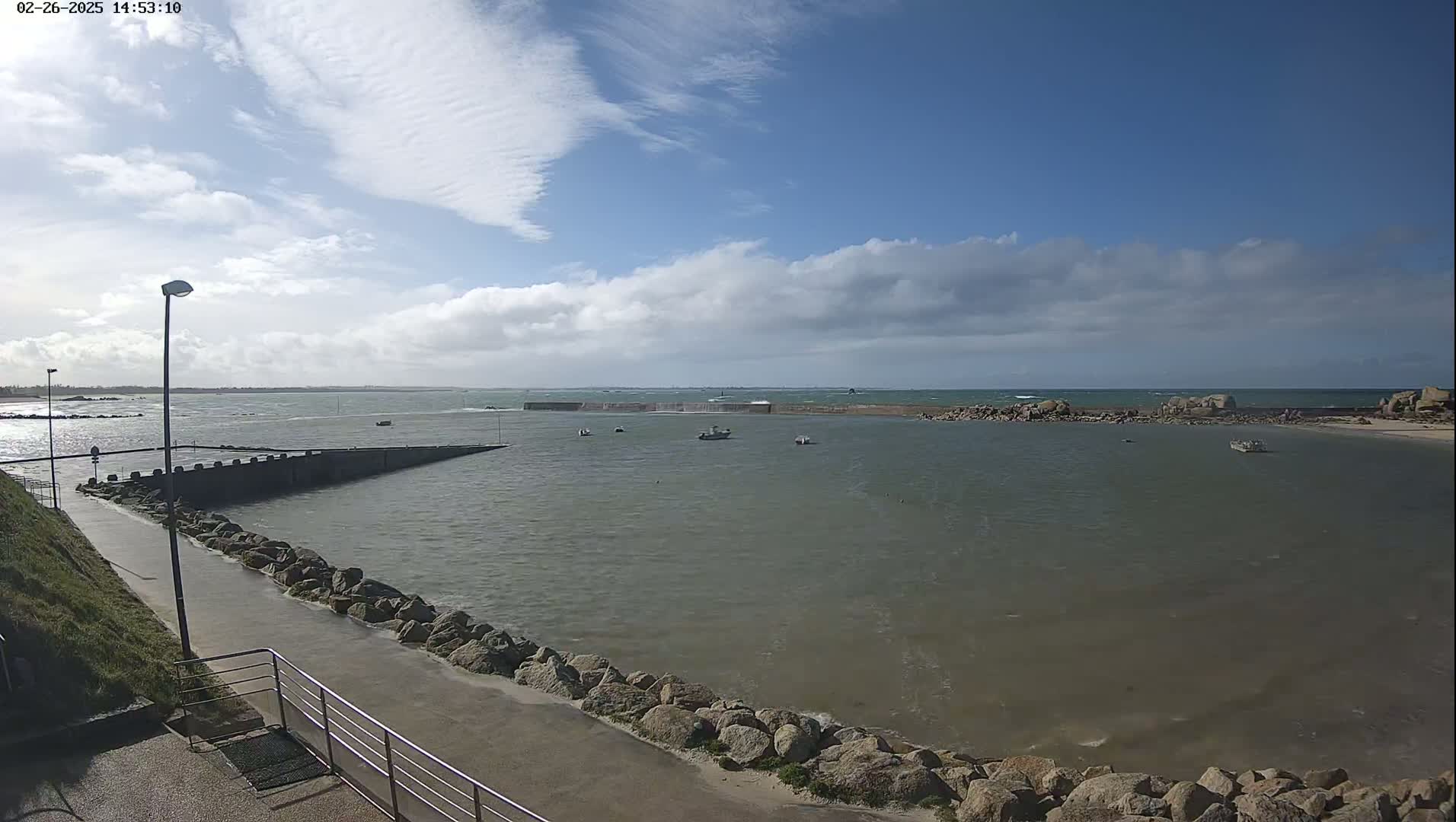 A partly cloudy day shows a calm harbor with a few small boats, a paved walkway, and rocky outcroppings leading to a larger body of water with small waves.