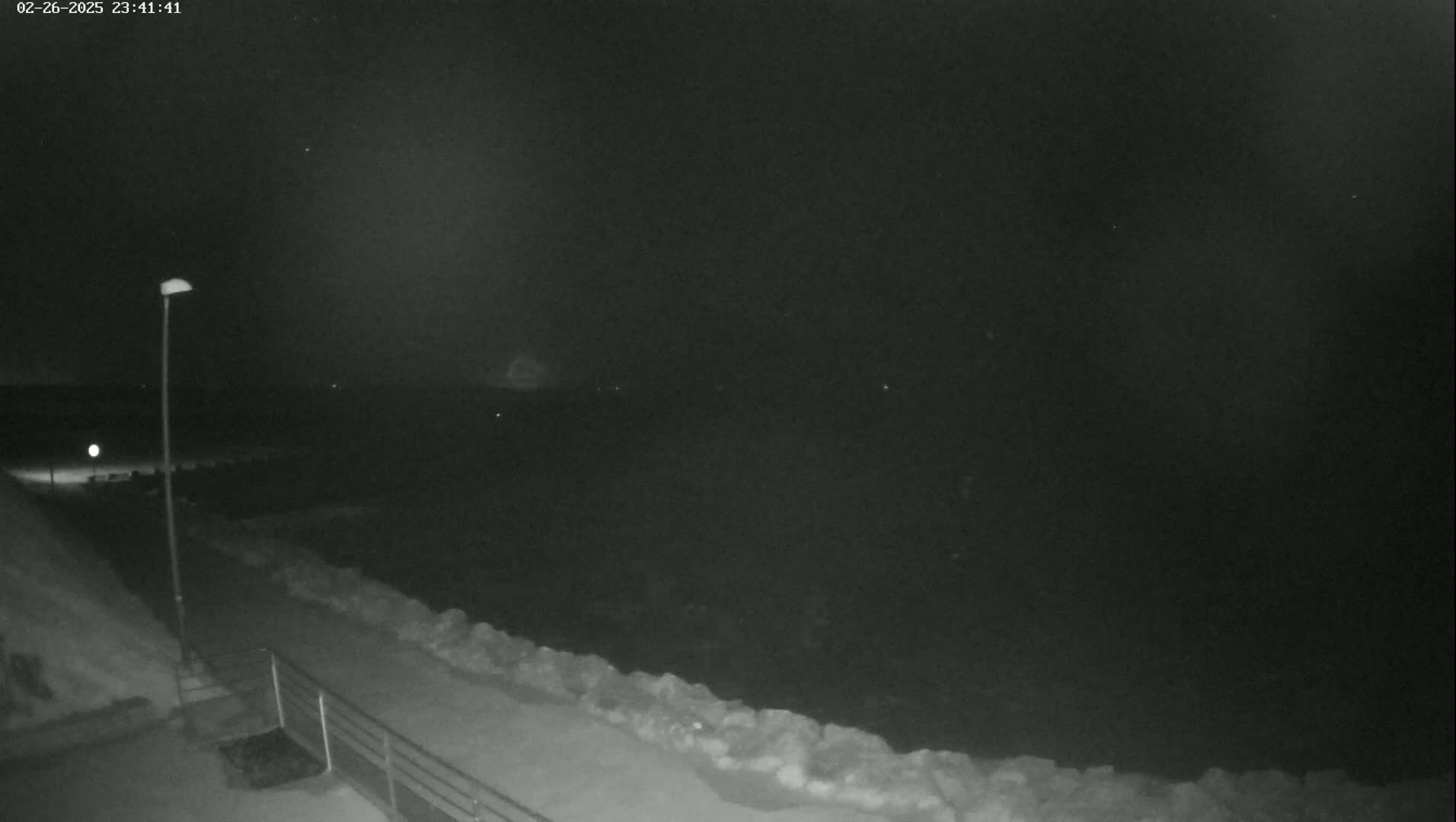A nighttime, snowy scene shows a dark body of water and a path along a snow-covered embankment, lit by a single lamppost.