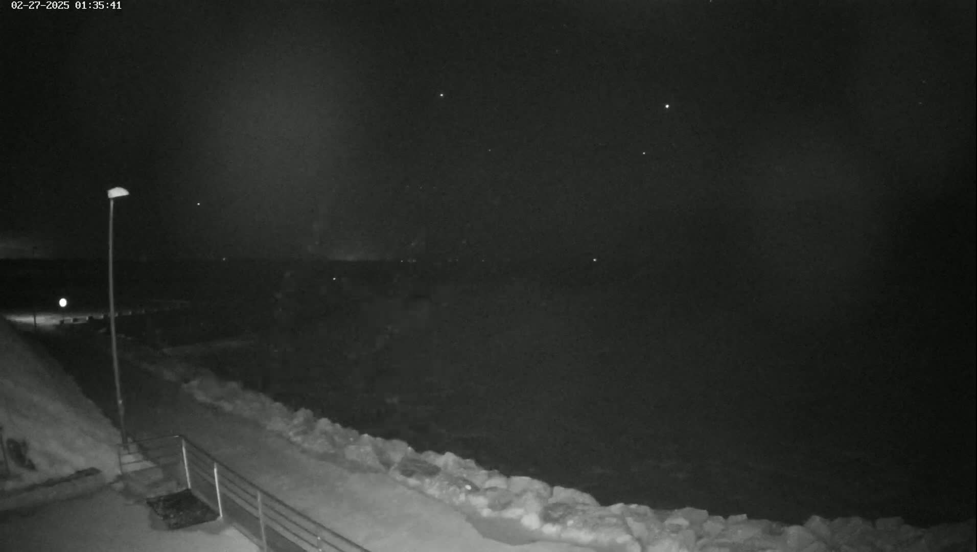 A nighttime, snow-covered walkway overlooks a dark, rough ocean under a mostly clear, starry sky.