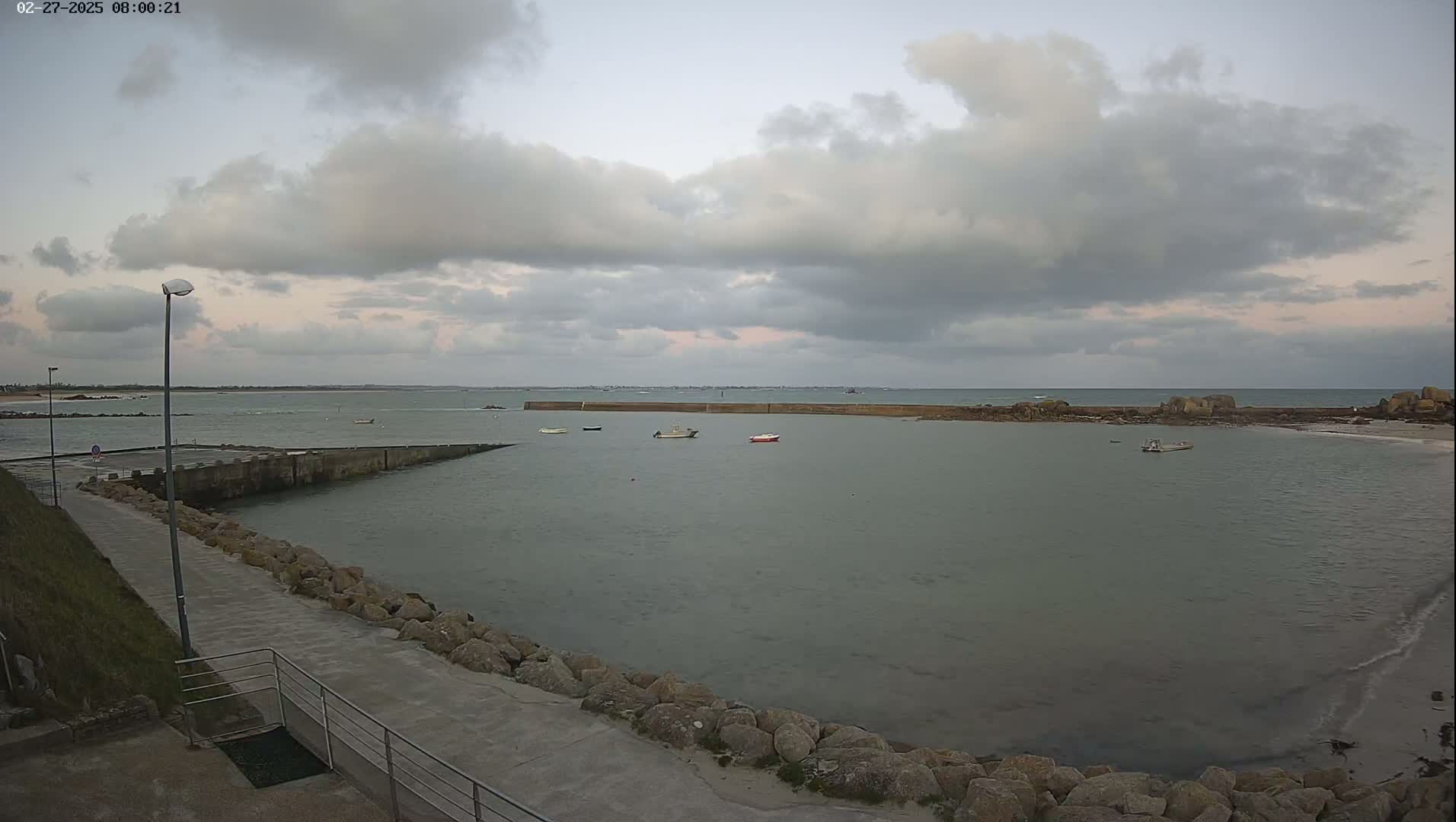 A calm, overcast day reveals a small harbor with several boats, a stone walkway, and a rocky shoreline extending to a calm sea under a cloudy sky.
