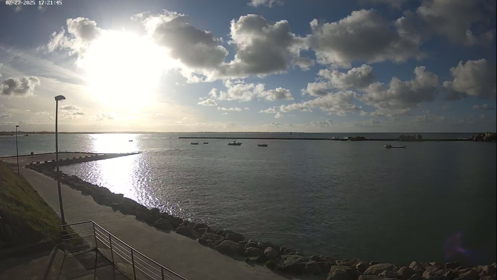 A partly sunny day with a few cumulus clouds over a calm harbor with several small boats, a paved walkway, and a rocky shoreline.
