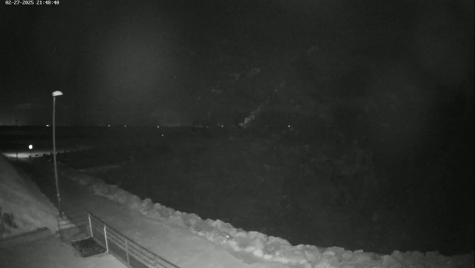 A nighttime view shows a snow-covered path next to a dark, icy body of water under a dark sky.