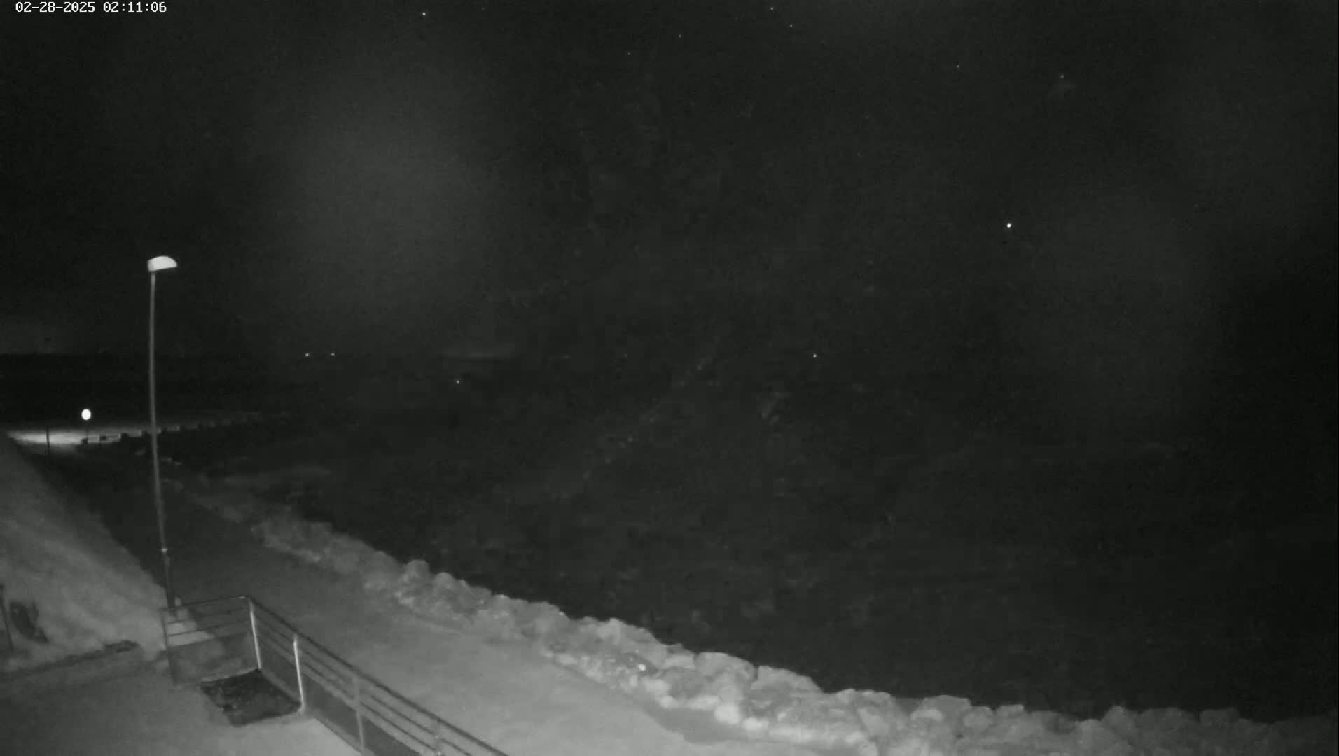 A nighttime, snow-covered scene shows a path leading to a dark body of water, lit by a single lamppost.