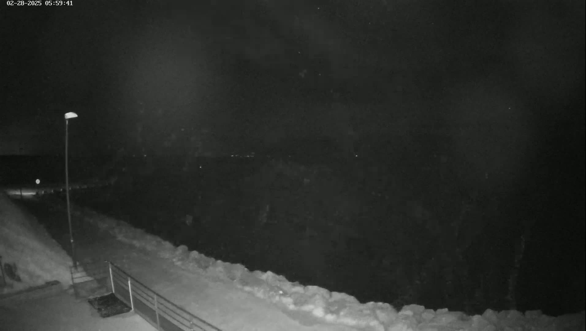A nighttime, snowy scene shows a snow-covered path leading to a metal railing overlooking a dark expanse under a lightly falling snow.
