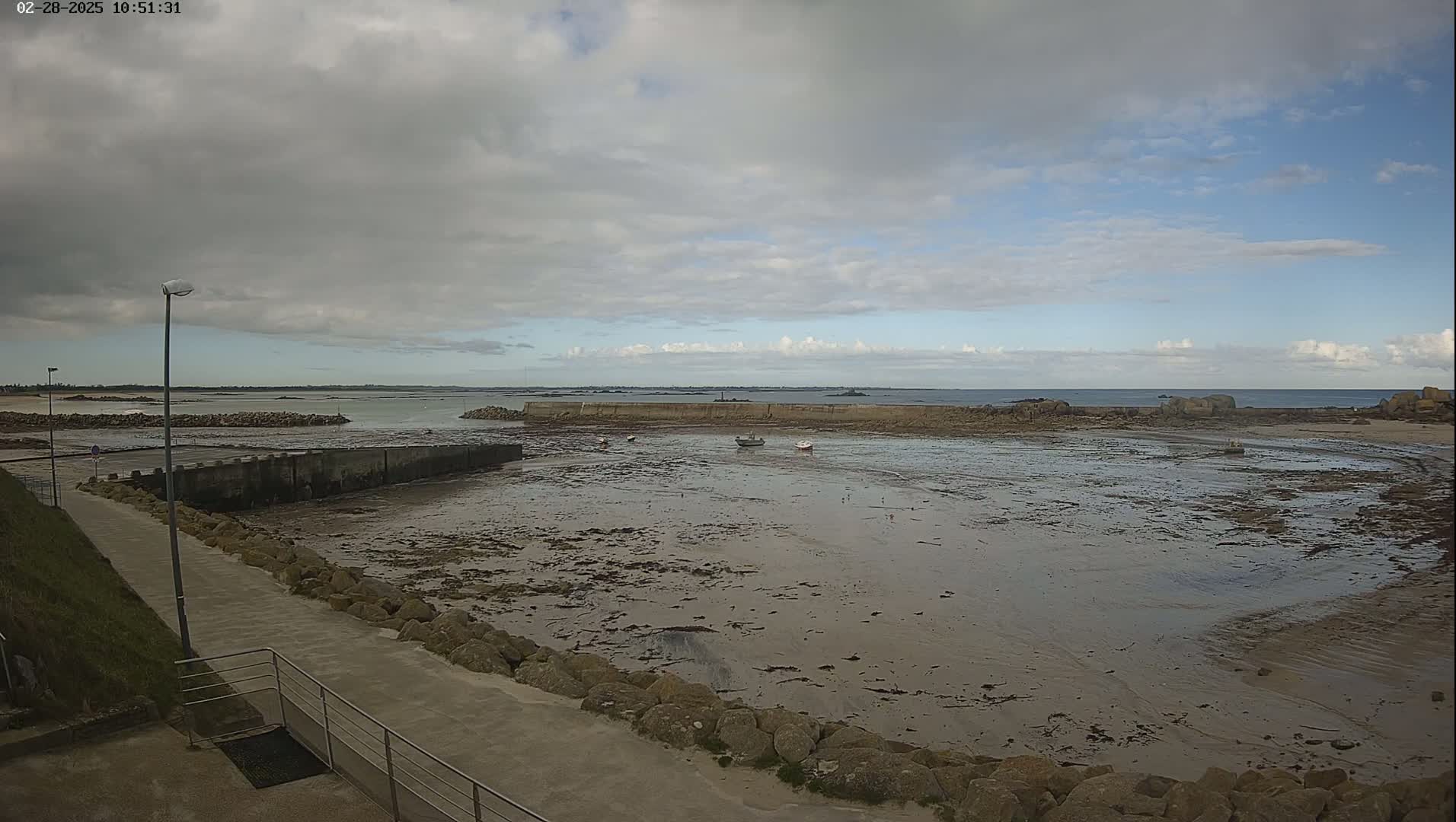 A mostly cloudy day reveals a low-tide harbor scene with several small boats, a concrete harbor wall, and a paved walkway.