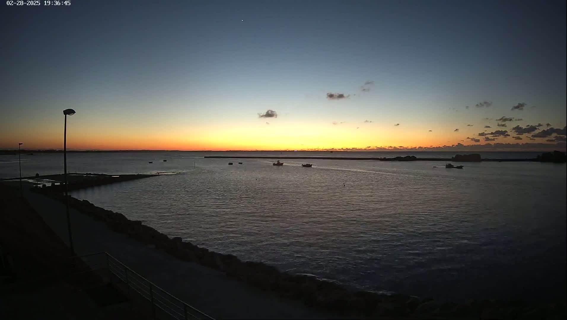 A calm body of water at sunset, with several small boats visible on the horizon, under a mostly clear sky.