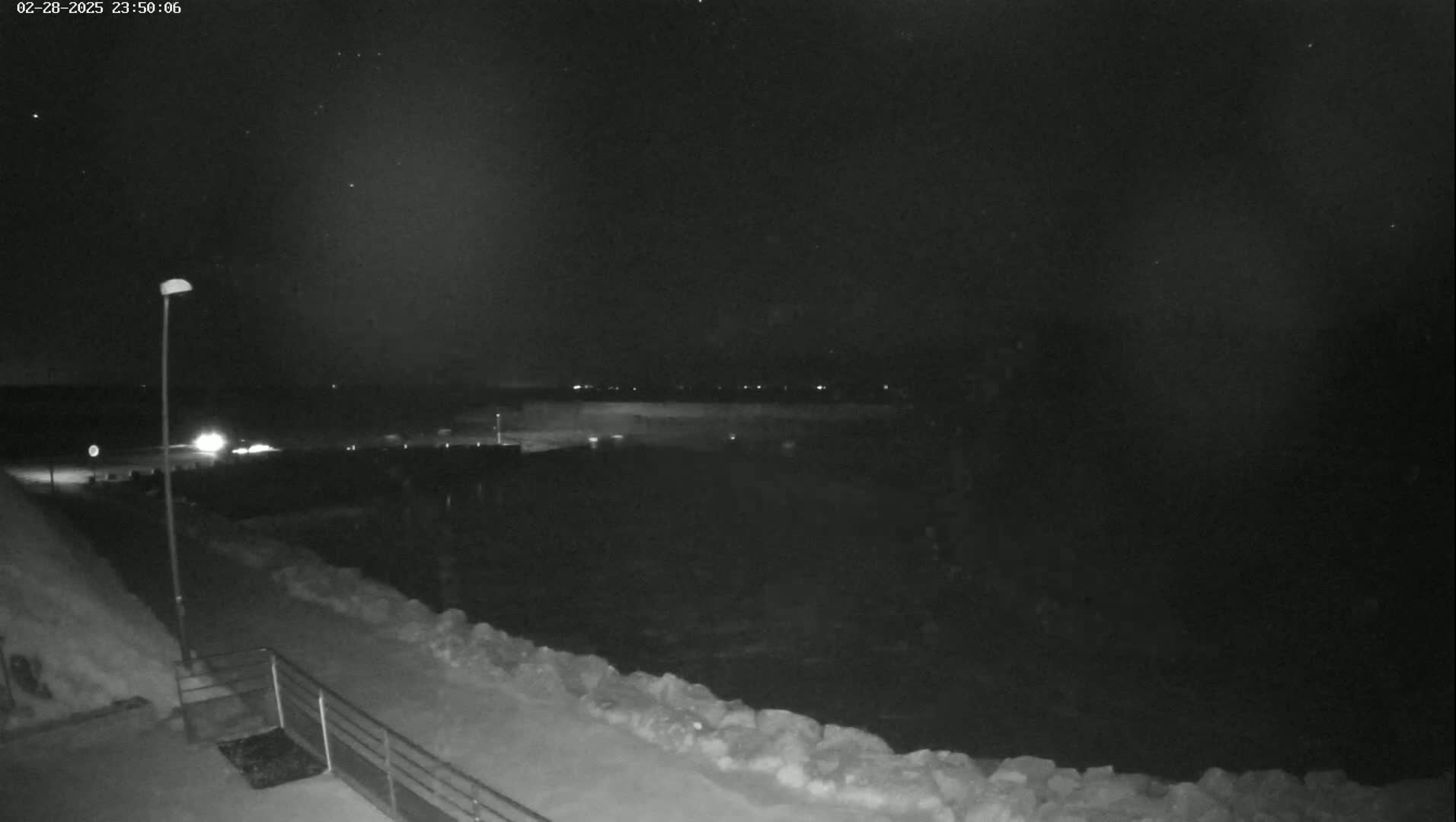 A nighttime view of a snow-covered waterfront, showing a dark body of water, a distant shoreline with faint lights, and a lamppost near a snow-covered walkway.