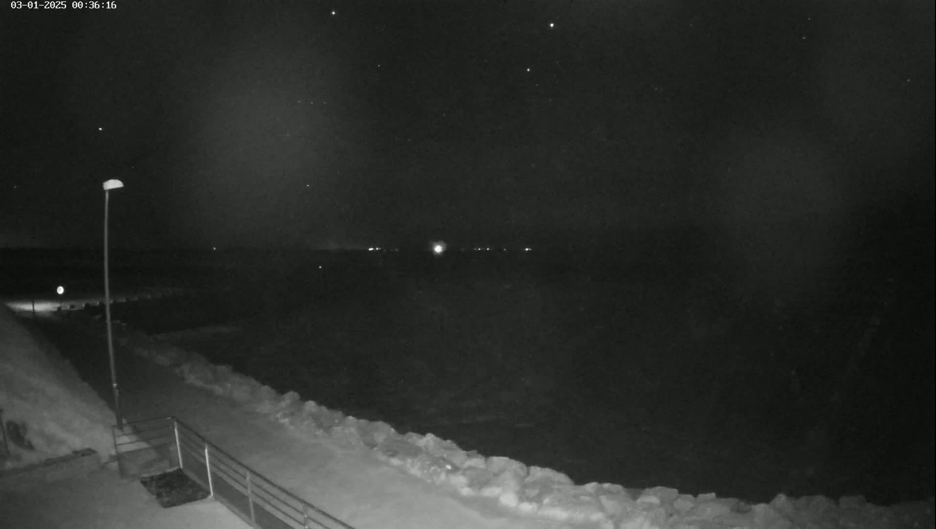 A nighttime view of a snow-covered shoreline with a single lamppost, under a dark, starry sky.