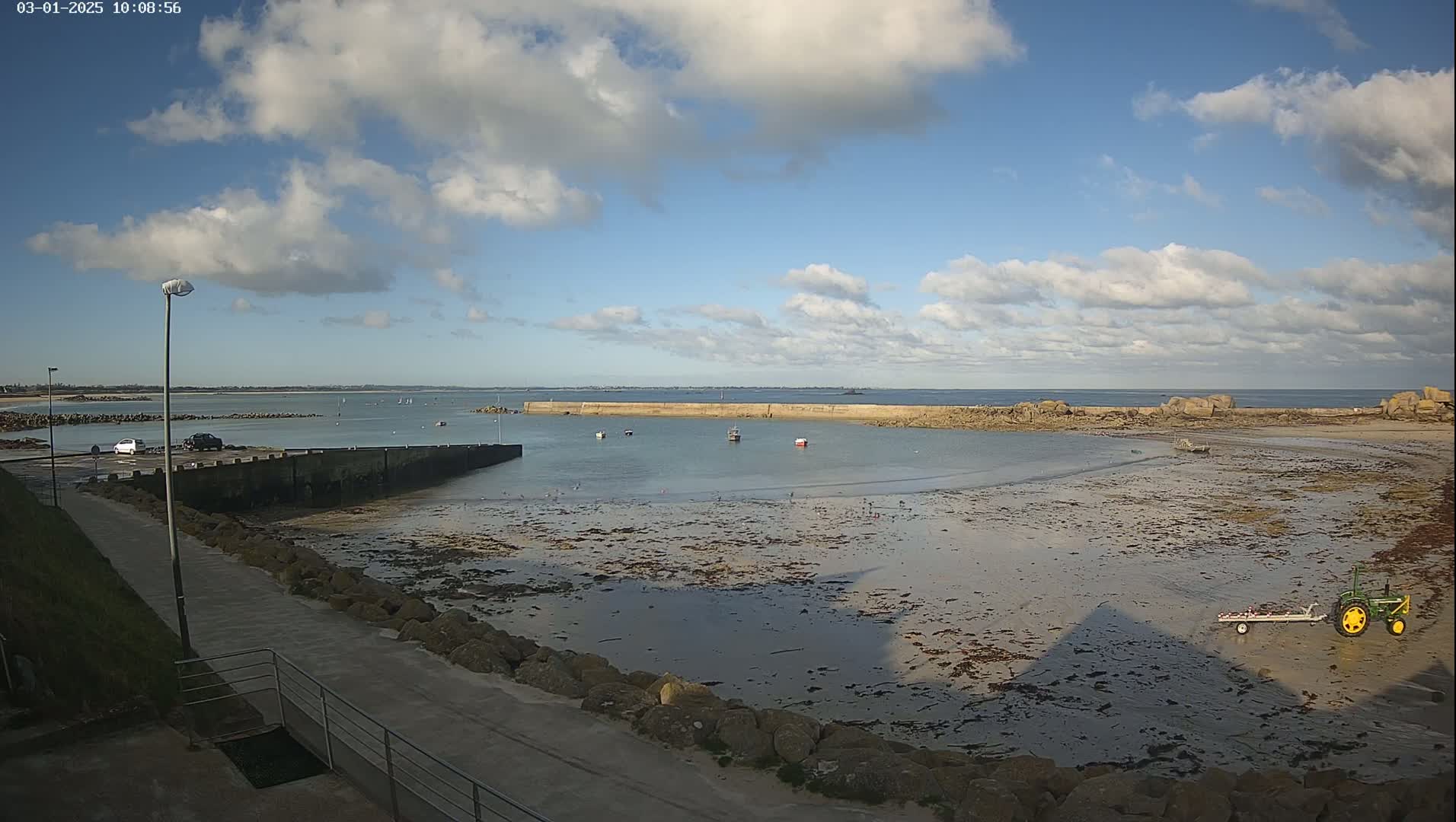 A partly cloudy day reveals a rocky, sandy beach with a small harbor containing several boats, a tractor and trailer on the shore, and cars parked nearby.