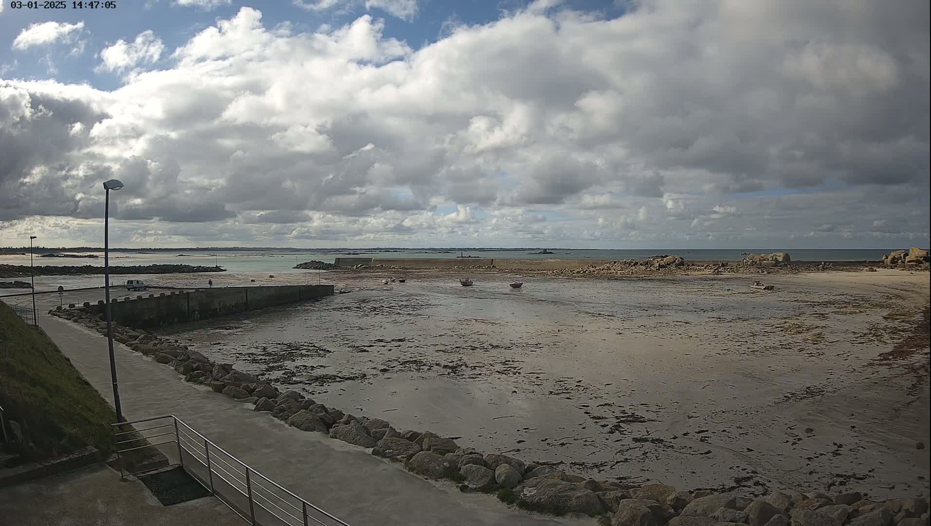 A mostly cloudy day reveals a low-tide harbor with several small boats, a walkway leading to it, and a calm ocean in the distance.