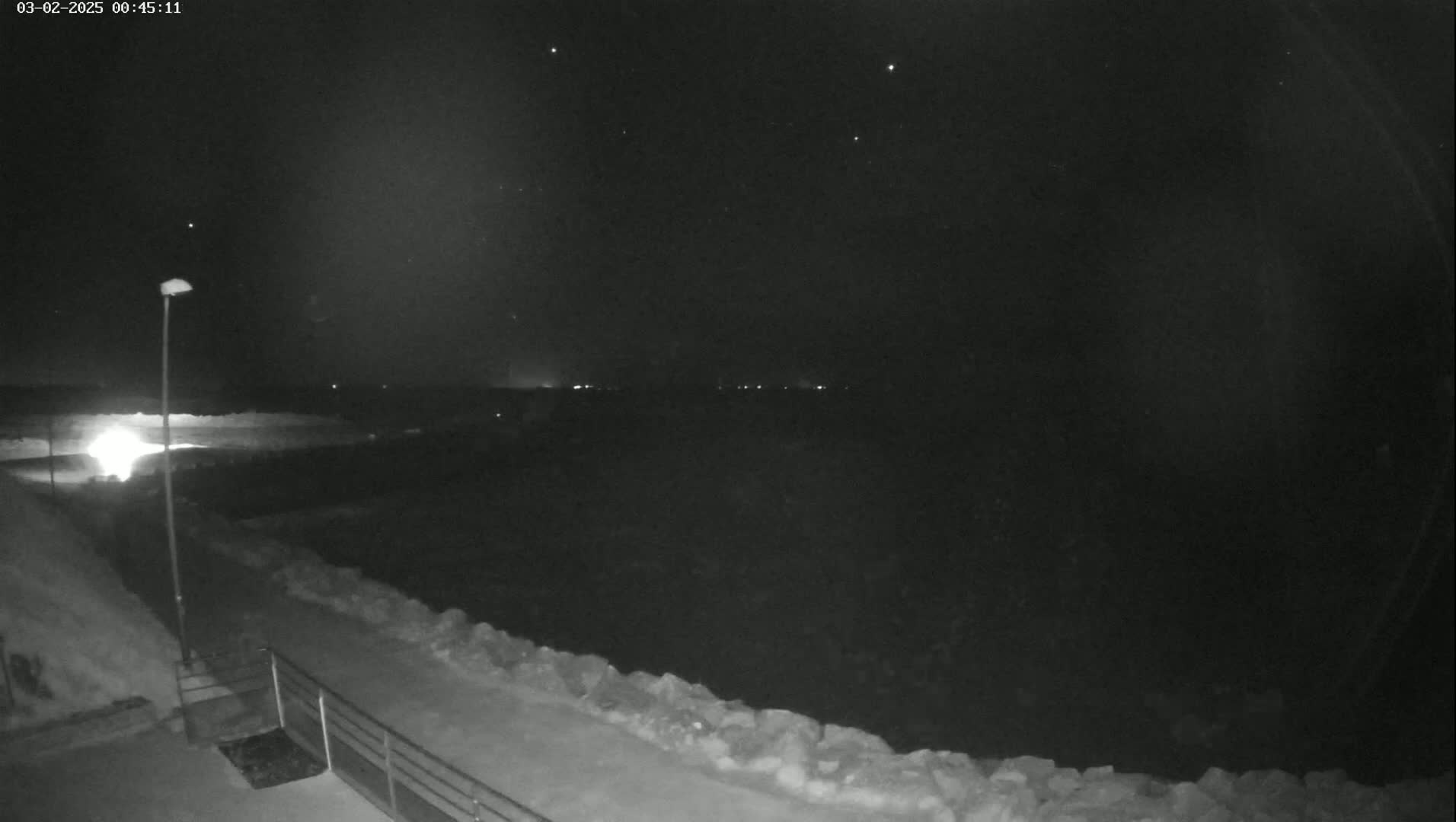 A nighttime view of a snow-covered landscape under a starry sky shows a path leading to a railing overlooking a dark body of water with distant lights.