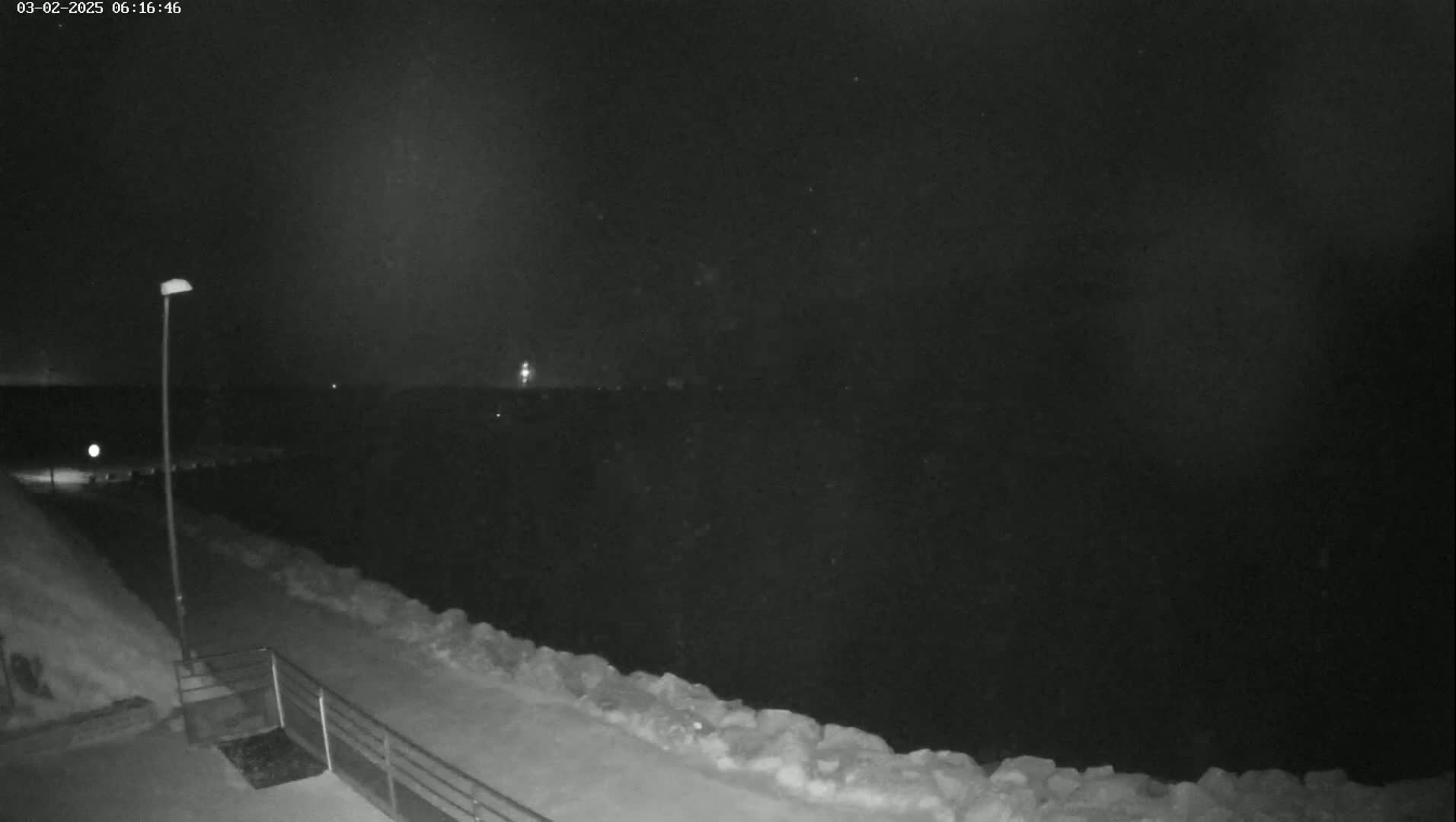 A nighttime, snow-covered path next to a body of water stretches towards a dark horizon under a clear sky.