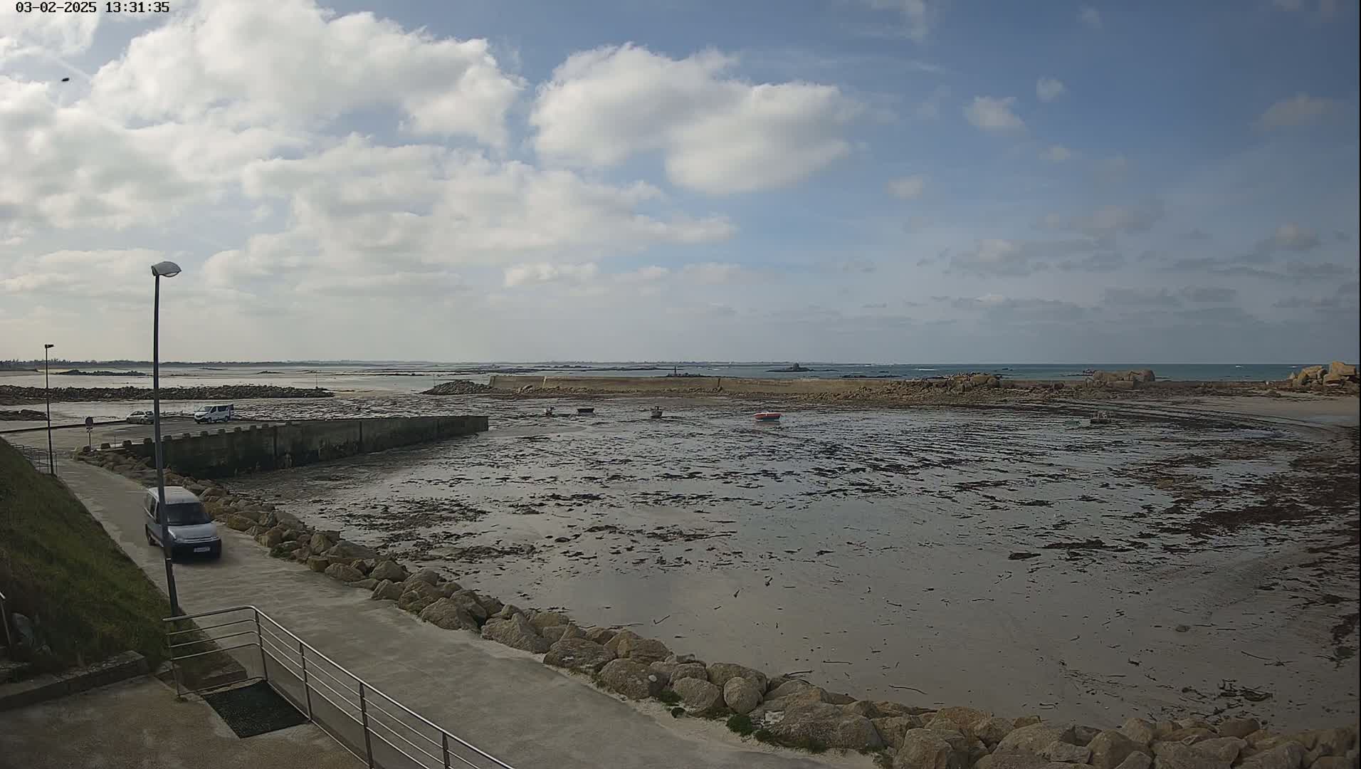 A partly cloudy day reveals a rocky, low-tide harbor with several small boats and a few cars parked nearby.
