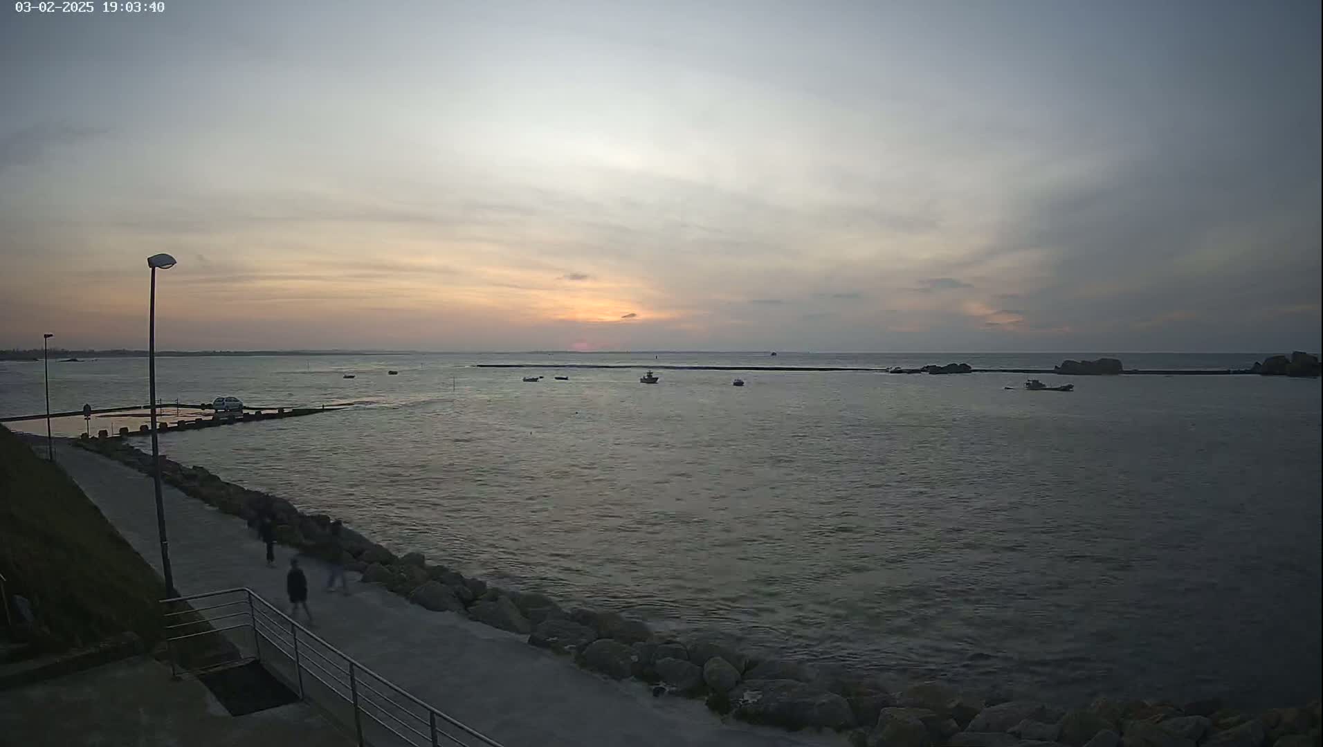 A calm body of water with several small boats at sunset, viewed from a paved walkway beside a rocky shoreline.