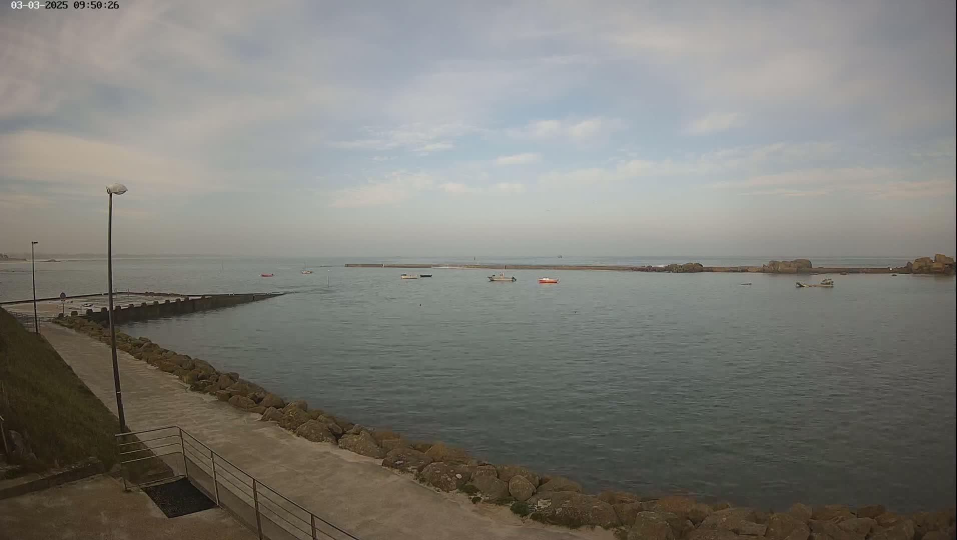 A calm body of water with several small boats is seen from a paved walkway next to a rocky shoreline under a partly cloudy sky.