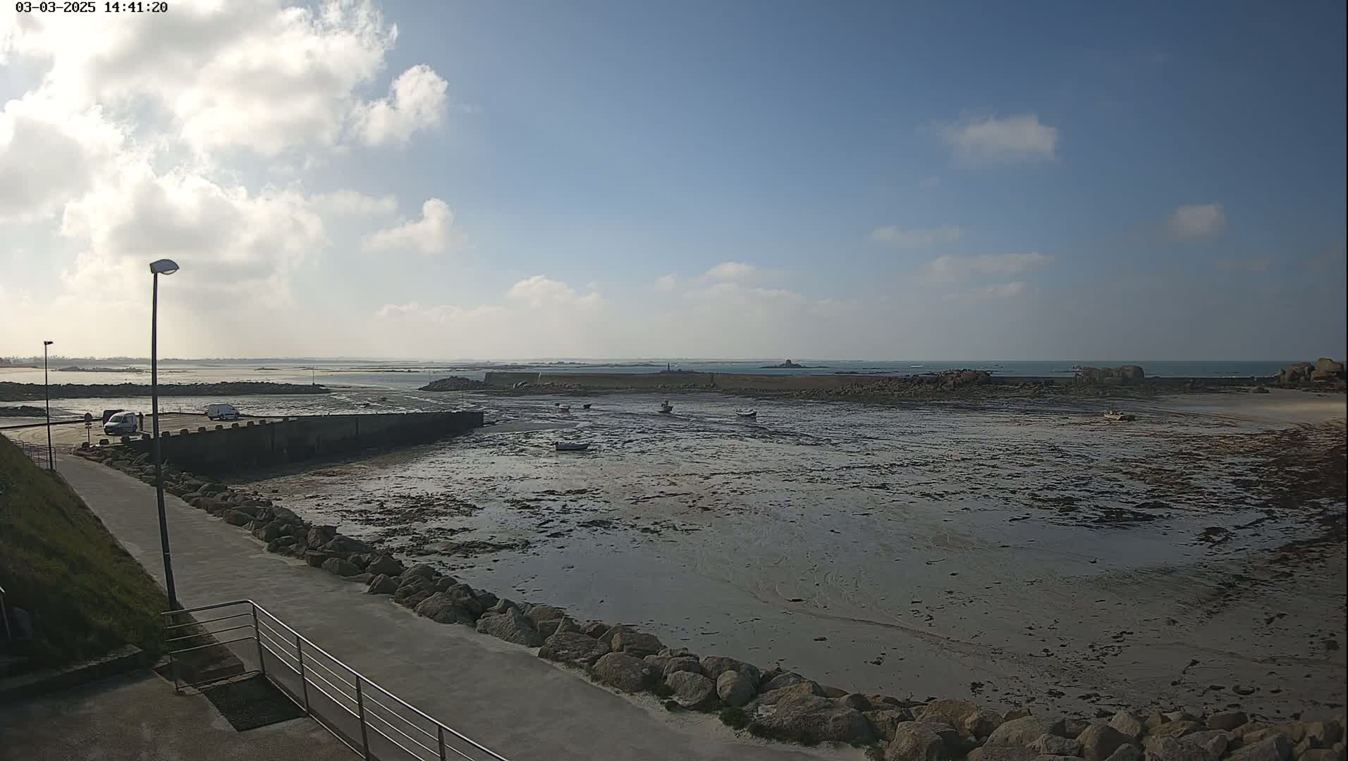 A partly cloudy day reveals a low-tide scene of a rocky harbor with several small boats, a walkway, and distant land under a mostly clear sky.