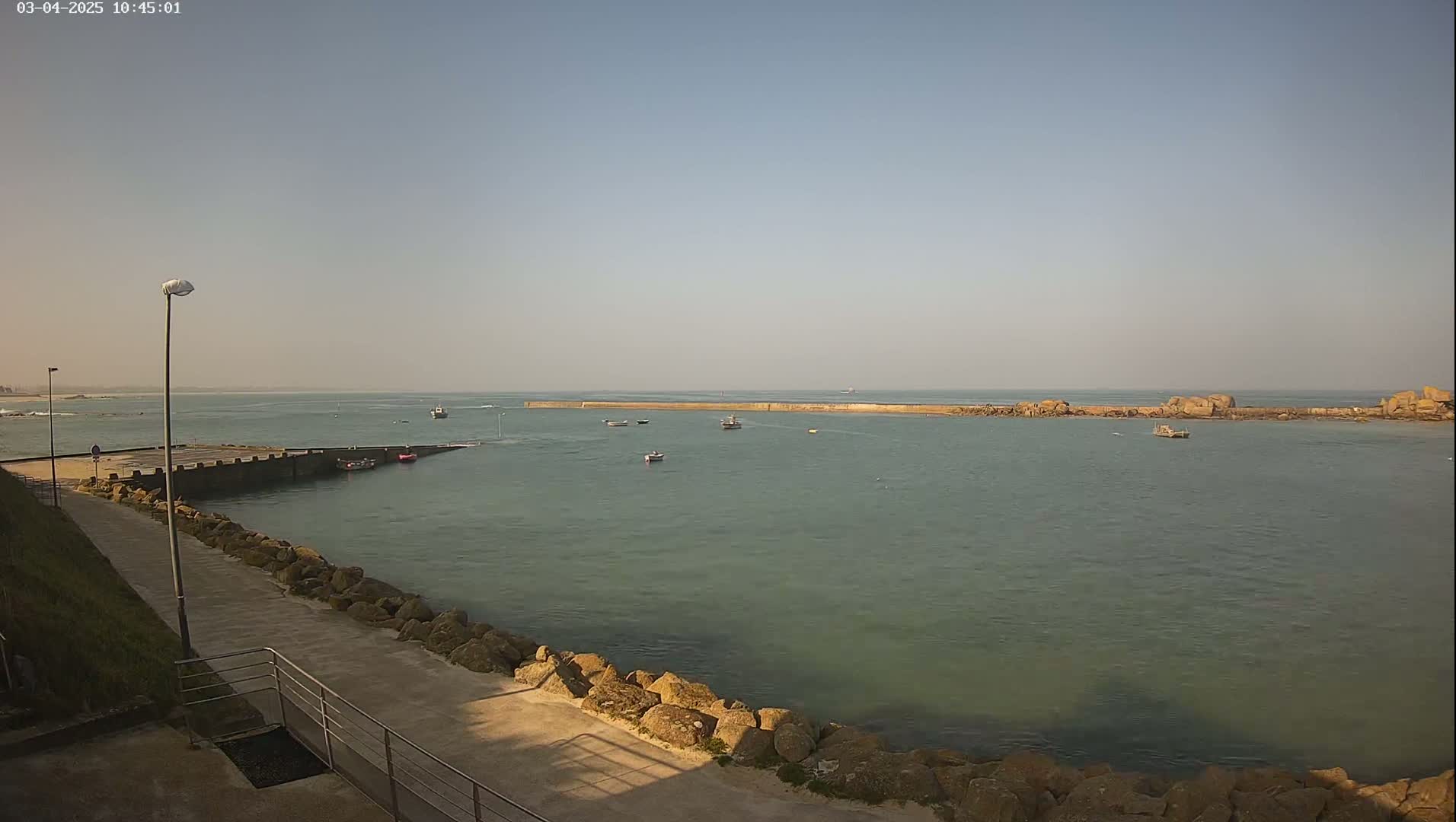 A calm harbor with several small boats is seen from a paved walkway next to a rocky shoreline under a clear sky.