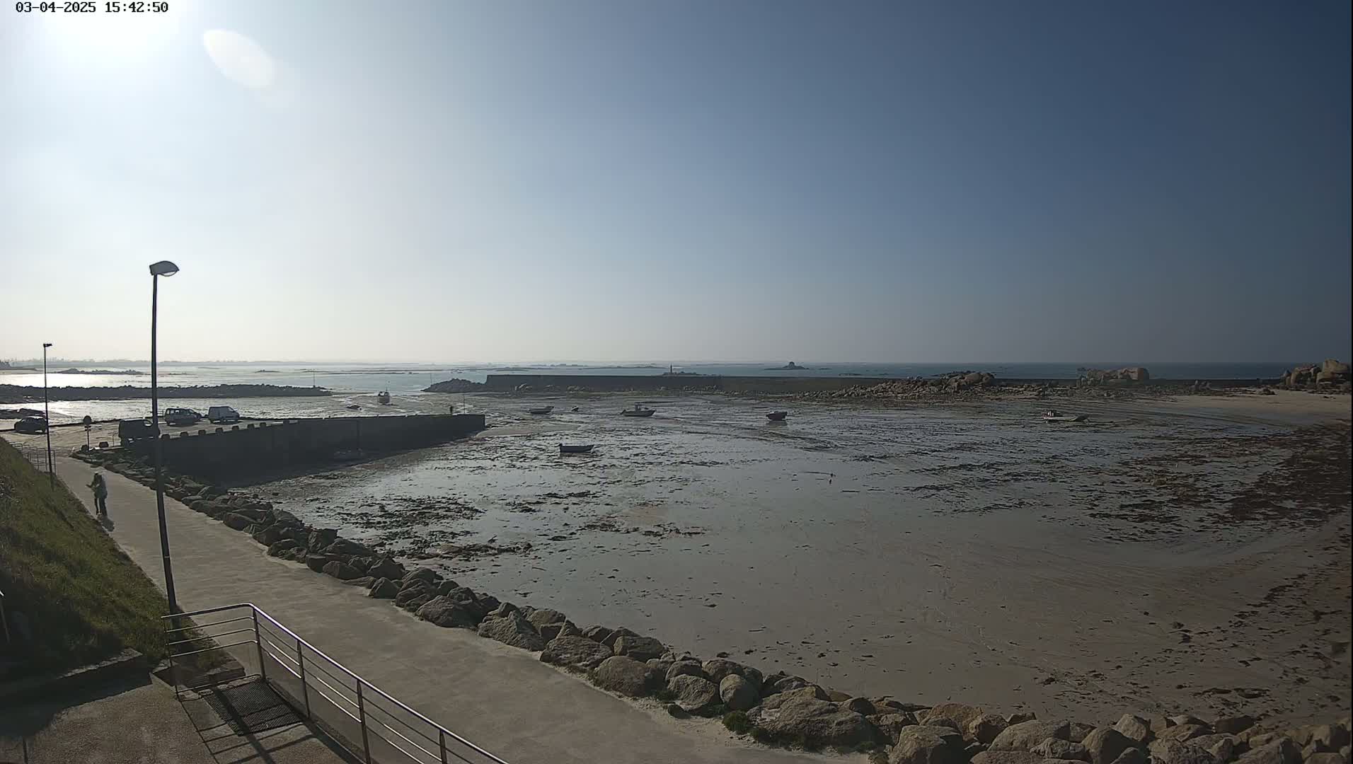 A mostly sunny day reveals a rocky shoreline with several small boats grounded on exposed mudflats at low tide, a walkway leading to a small harbor, and distant ocean views.