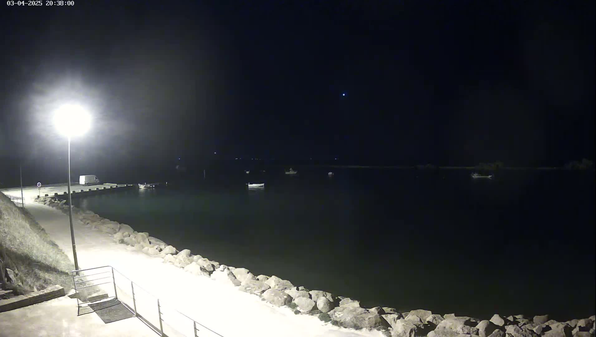 A nighttime view of a calm body of water with several boats, a lit walkway, and a van parked near a dock under a dark, clear sky.