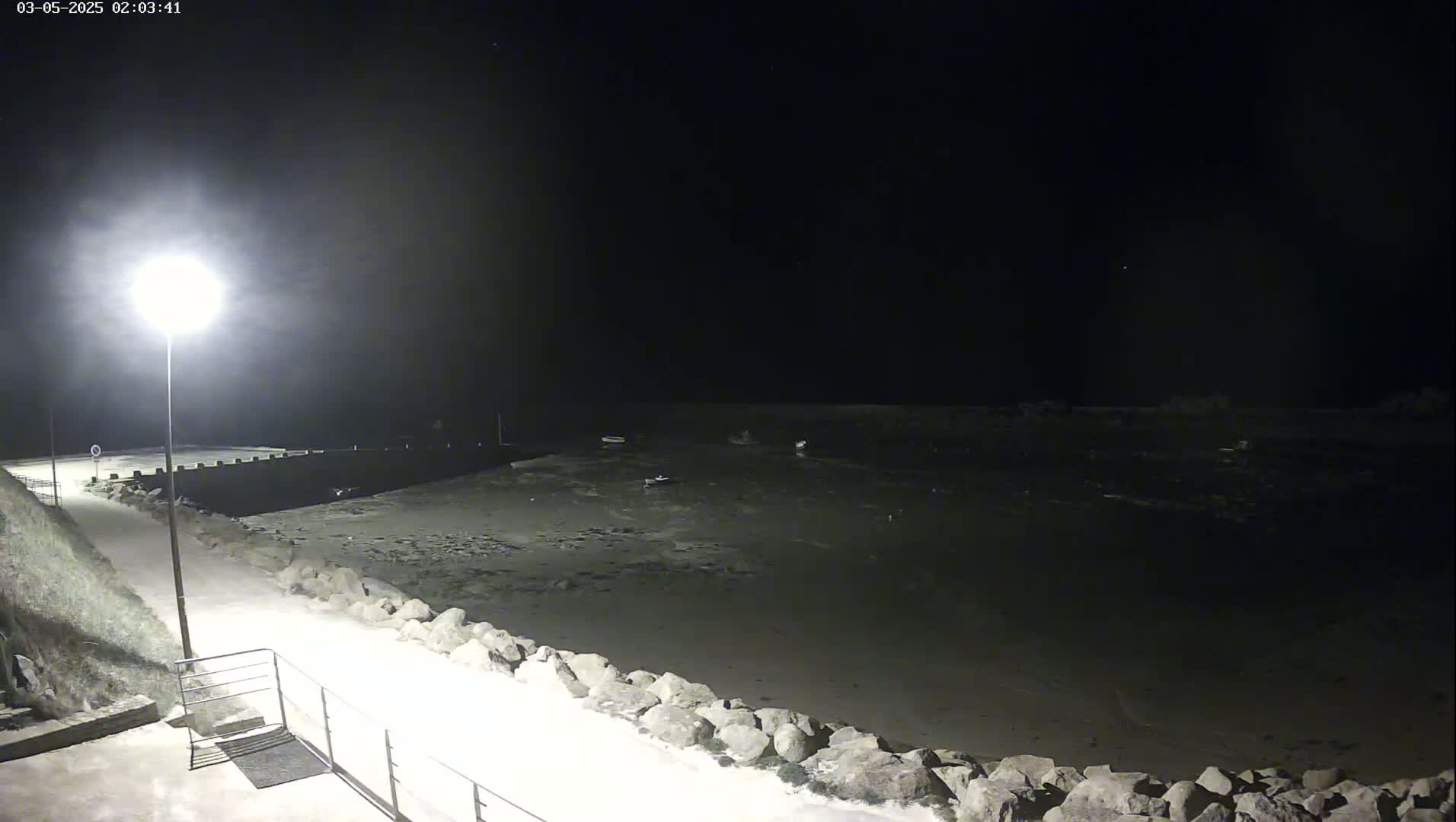 A nighttime view of a calm, dark body of water next to a paved walkway lined with rocks, illuminated by a single lamppost.