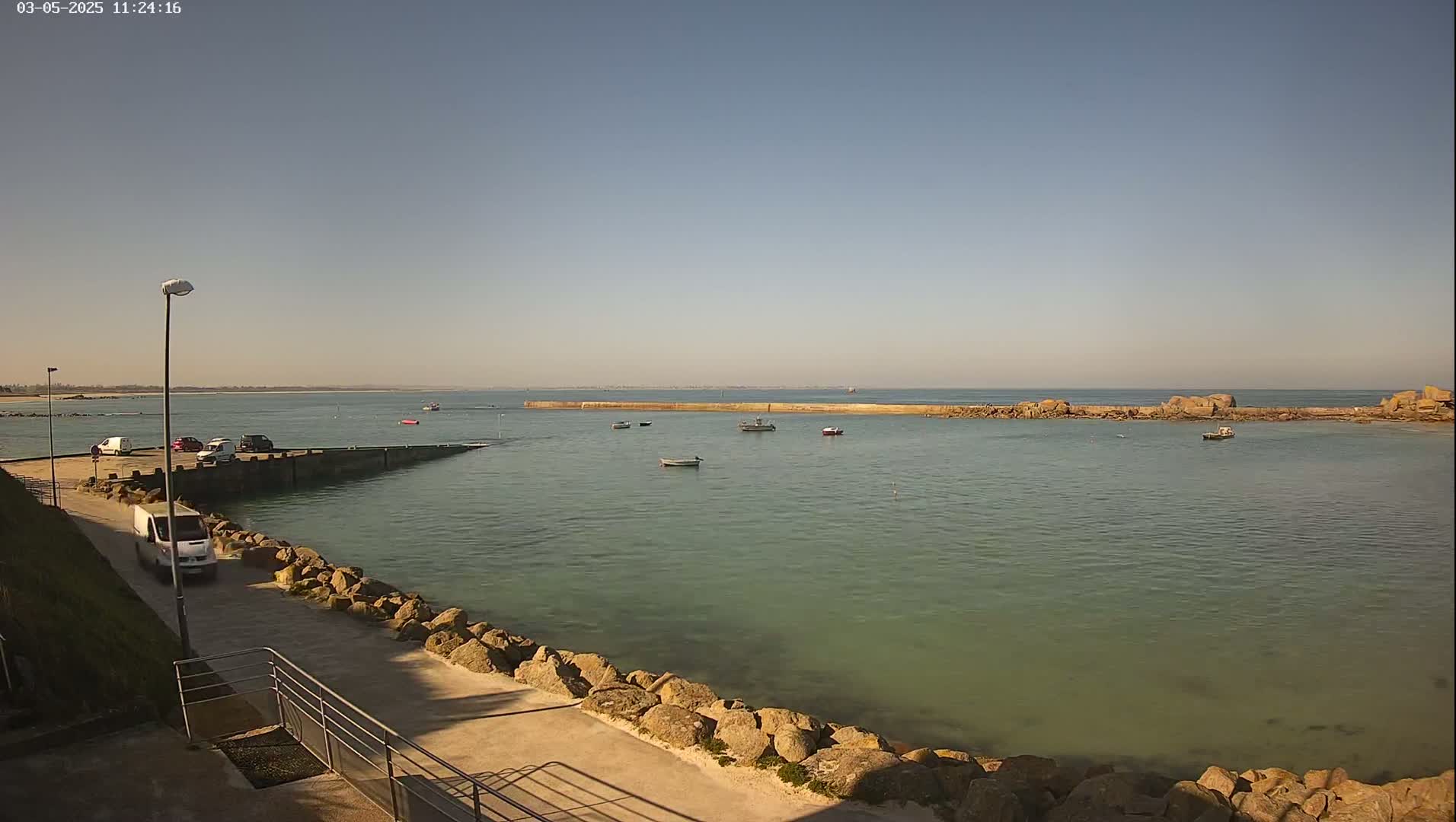 A calm, sunny day reveals a harbor with several small boats, a breakwater, and a paved walkway lined with rocks.