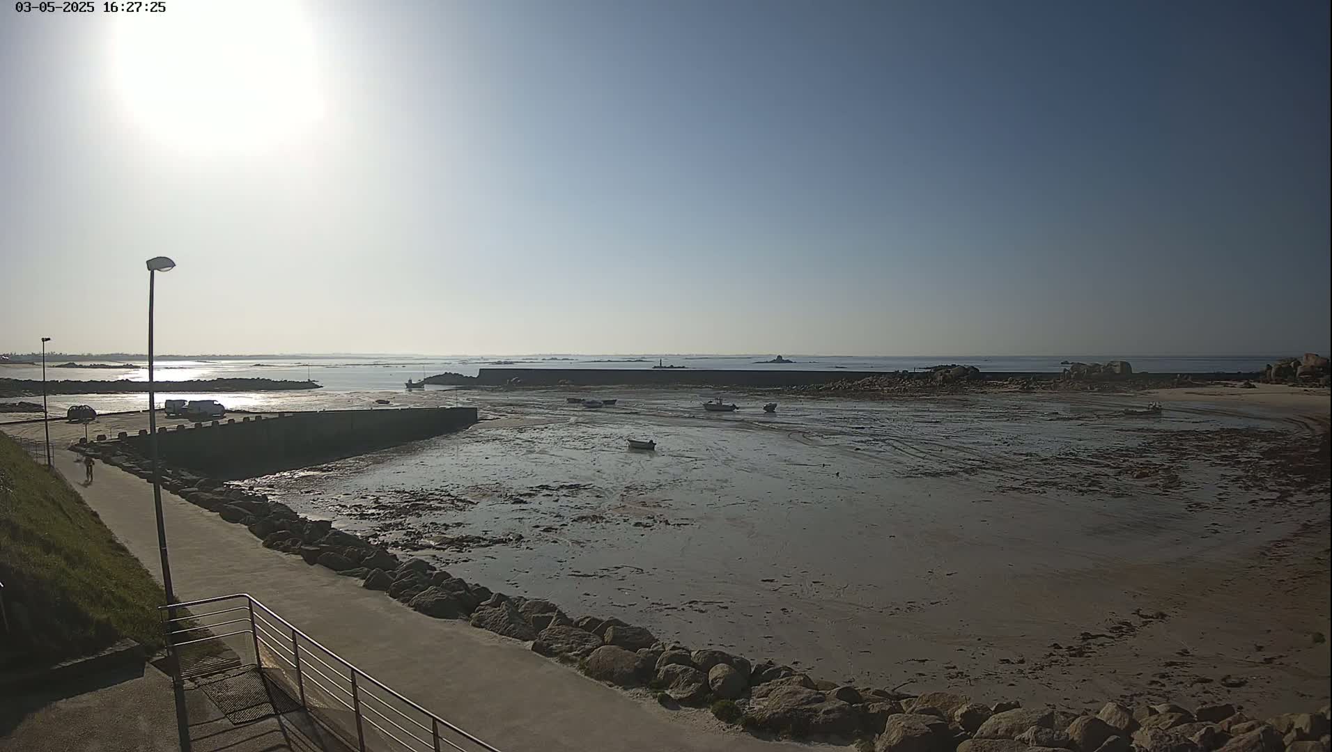 A mostly sunny day reveals a harbor with several small boats resting on exposed mudflats at low tide, surrounded by a low stone wall and rocky shoreline.
