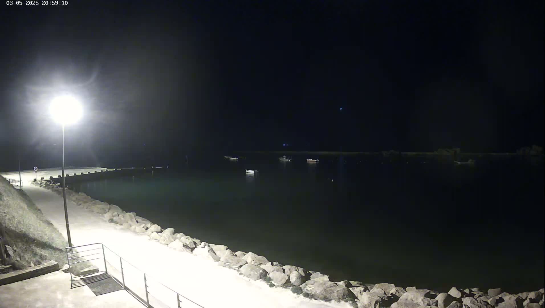 A nighttime view of a calm body of water with several small boats moored, a lit pathway leading to a small pier, and a rocky shoreline.