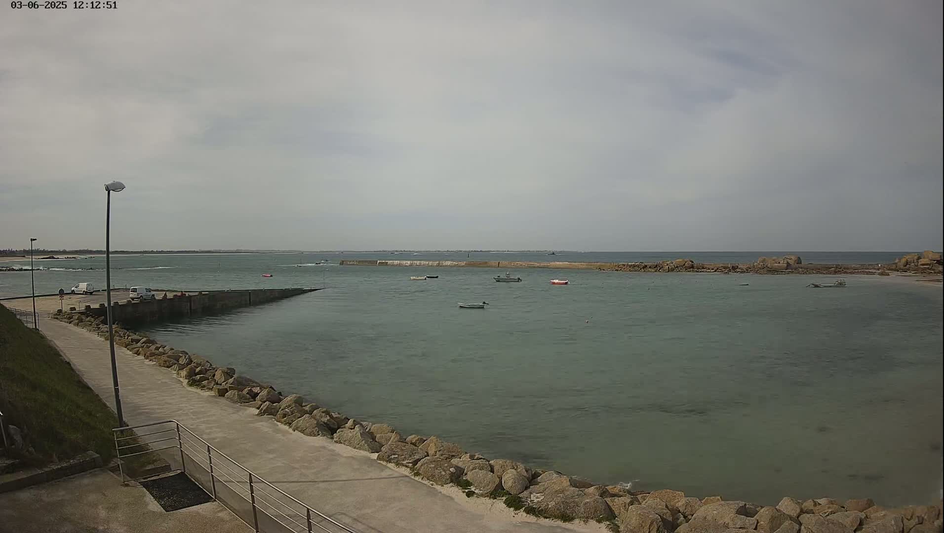 A calm, overcast day reveals a harbor with several small boats, a paved walkway lined with rocks, and a distant coastline.