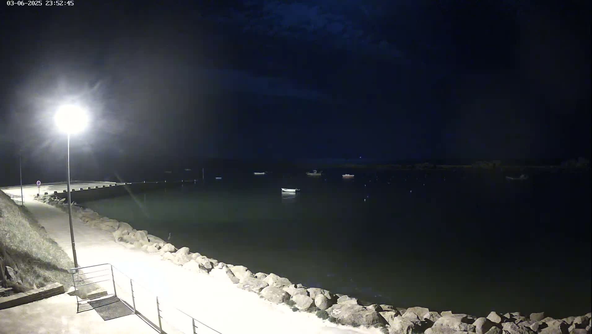 A nighttime view of a calm harbor with several small boats moored, illuminated by a single lamppost on a stone walkway.