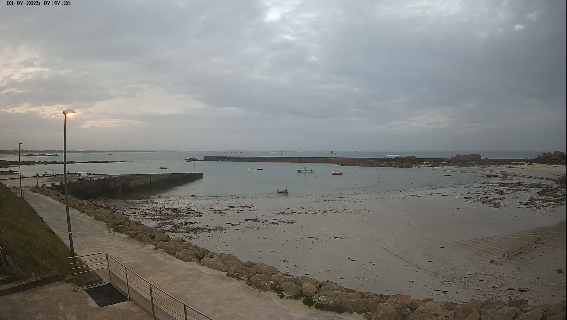 A calm, overcast day reveals a small harbor with several boats, a rocky shoreline, and a paved walkway leading to the water.