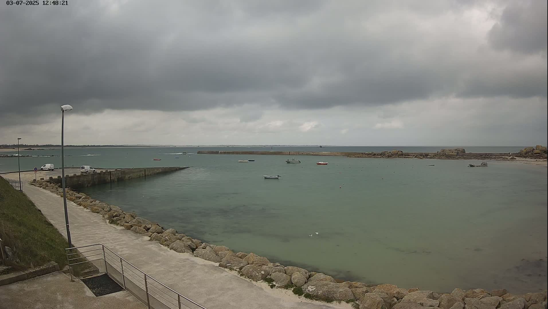 A calm, overcast day reveals a harbor with several small boats, a stone walkway, and a few parked vehicles.