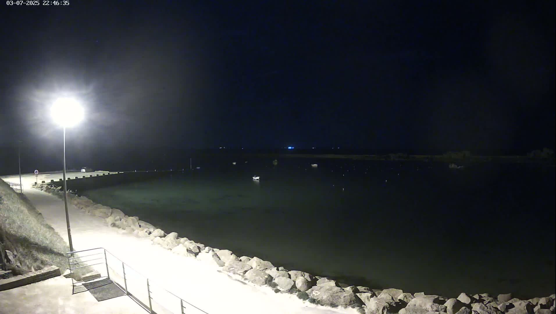 A nighttime view of a calm body of water, partially illuminated by a single lamppost on a paved walkway lined with large rocks, shows several small boats moored in the distance.