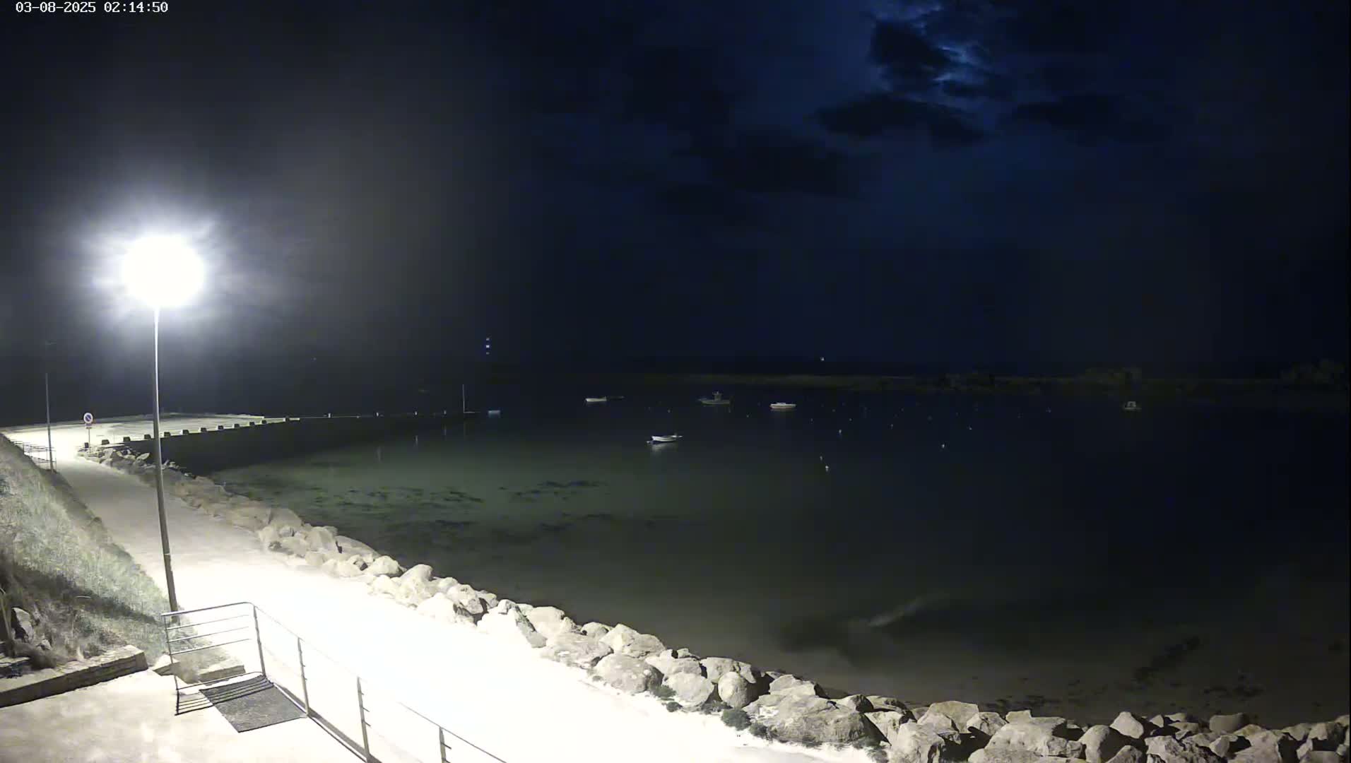 A moonlit night scene shows a calm body of water with several small boats, a paved walkway lined with rocks leading to a bright lamppost, and a dark, cloudy sky.