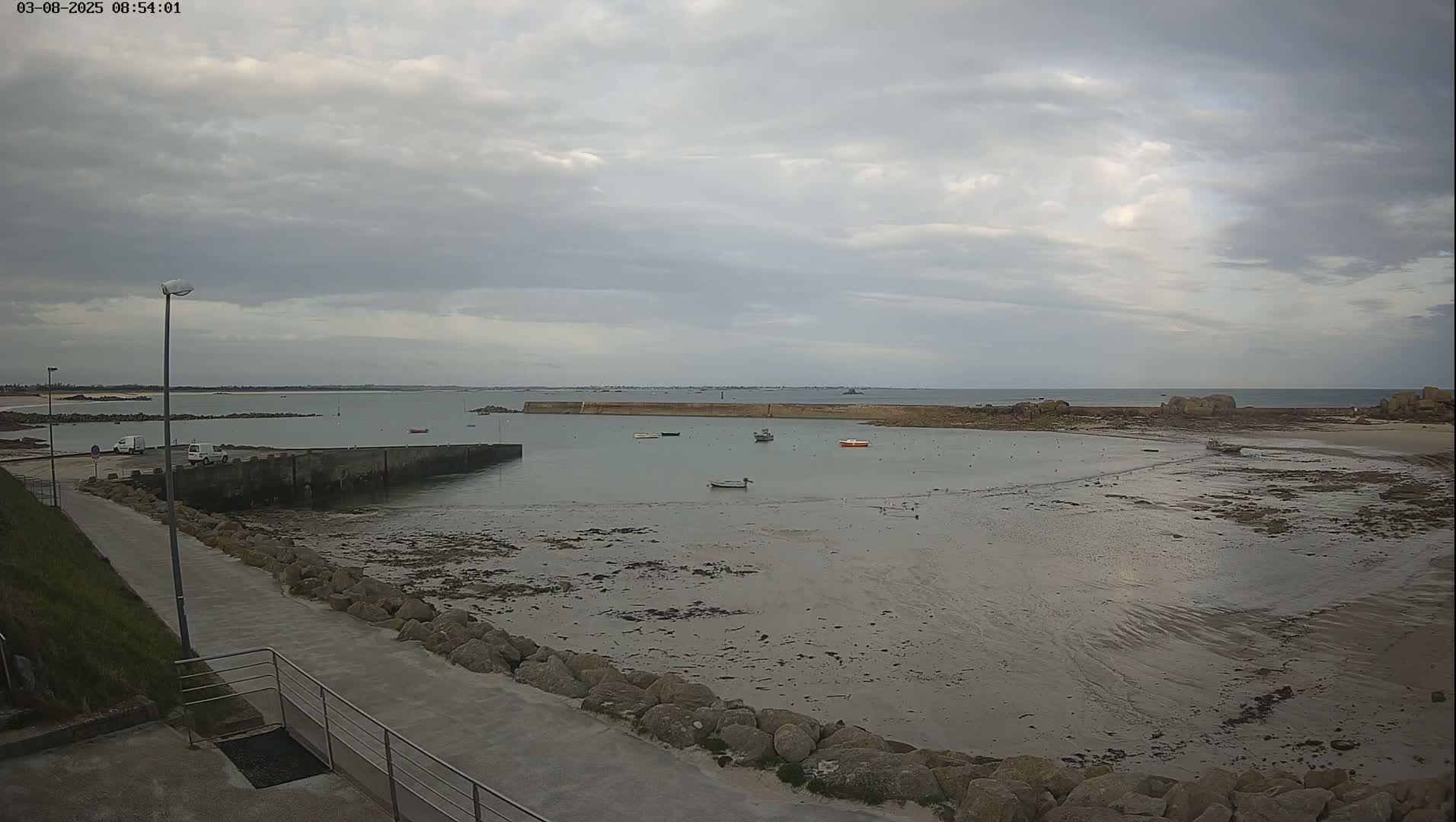A mostly cloudy day reveals a harbor with several small boats at low tide, a paved walkway, and a rocky shoreline.
