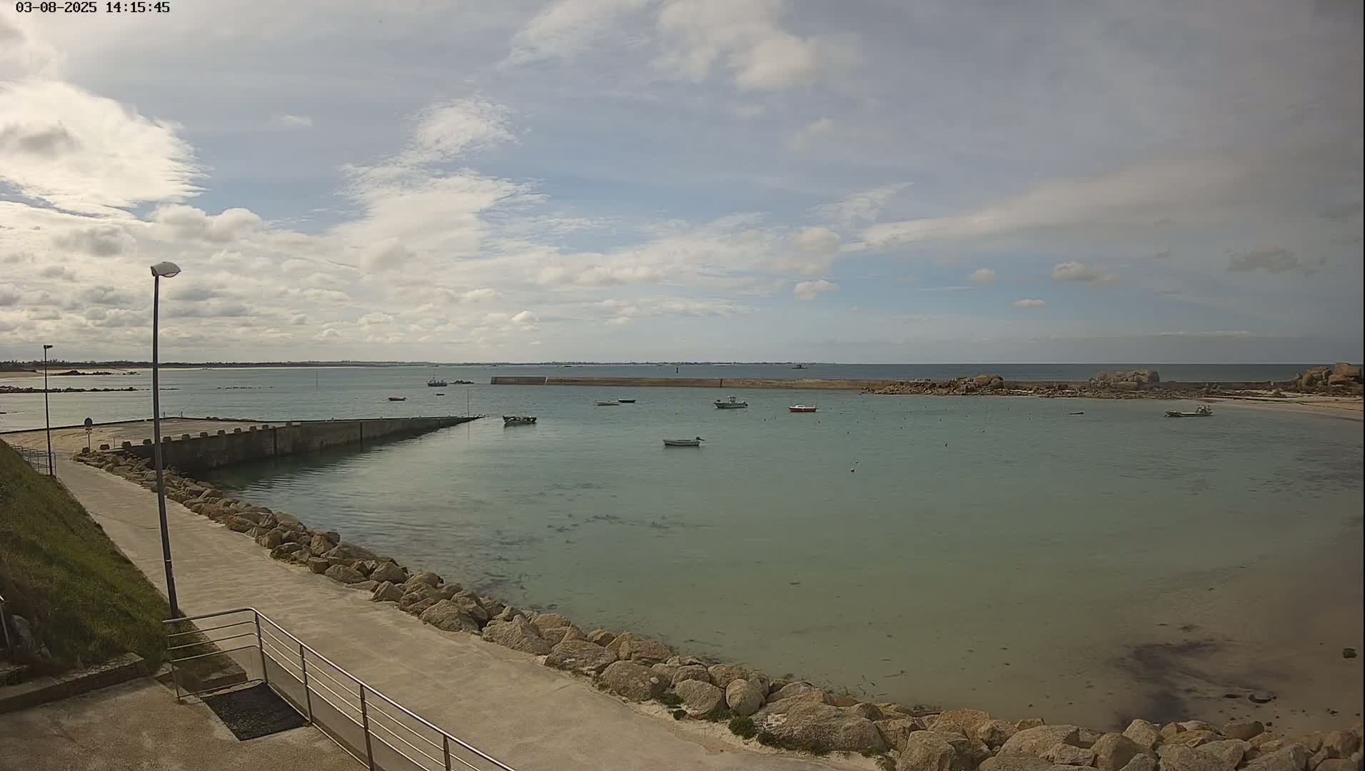 A calm, partly cloudy day reveals a small harbor with several boats moored, a paved walkway lined with rocks leading to the water's edge, and a distant coastline.